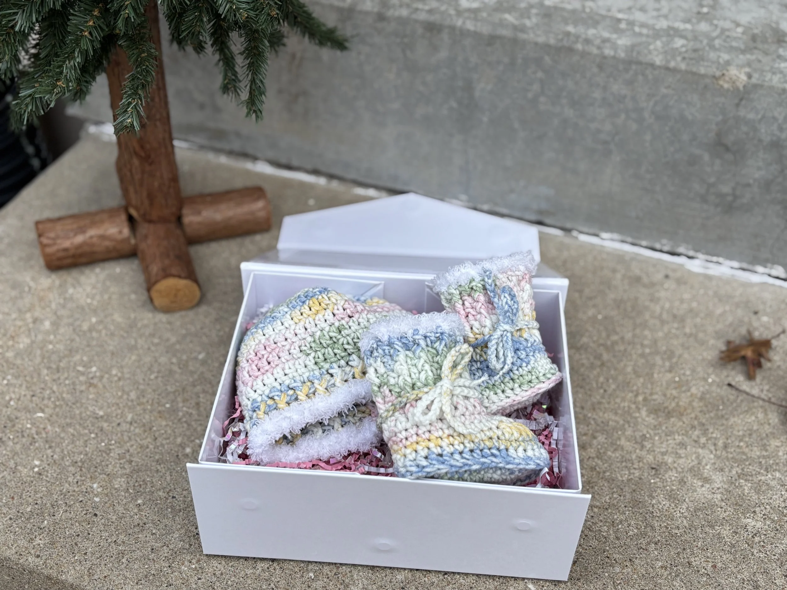 A pair of multicolored crocheted infant booties and a matching hat inside a white gift box placed on a concrete surface, next to a small wooden Christmas tree.