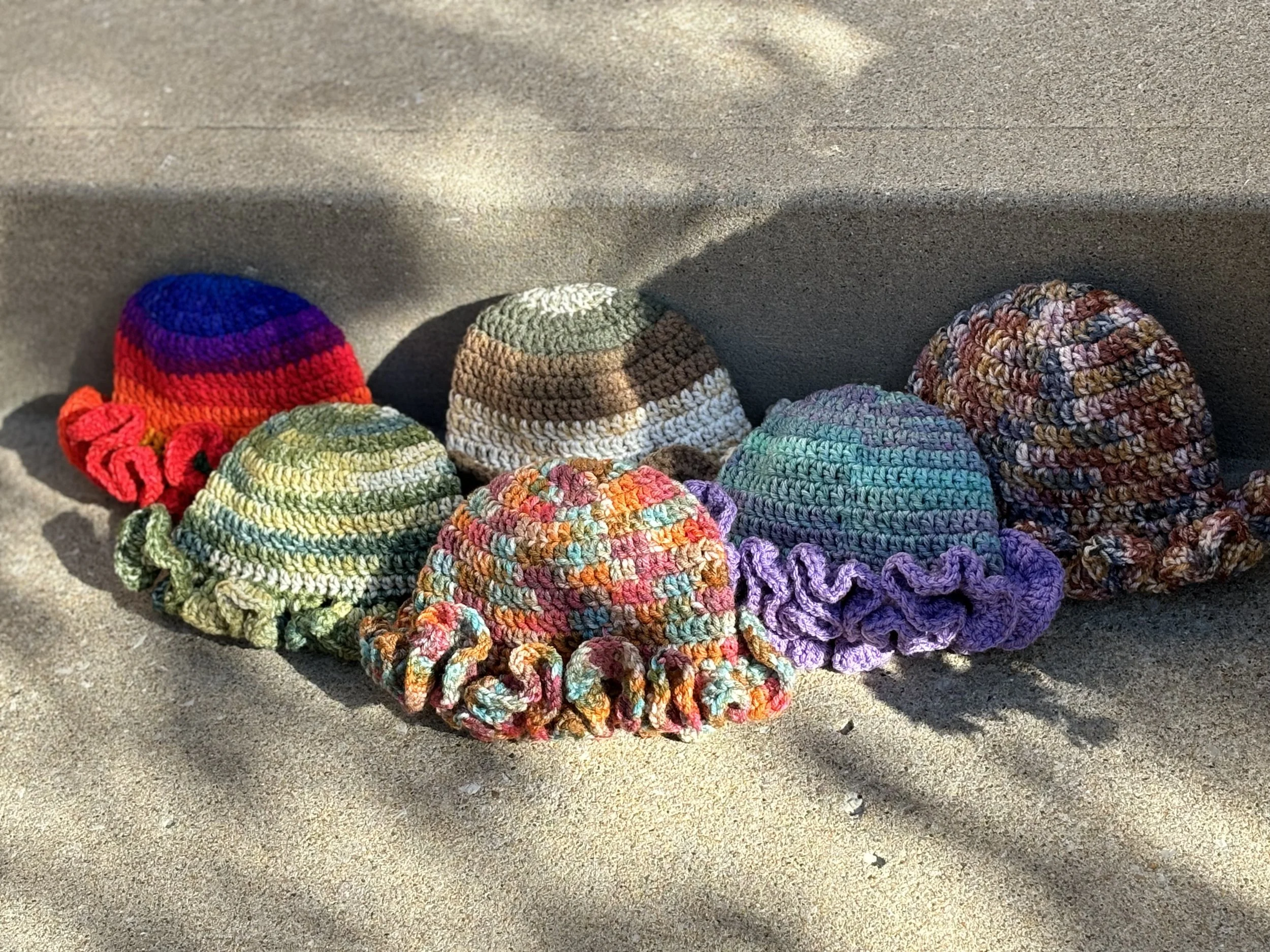 Six colorful crocheted hats with ruffled brims, displayed on a concrete surface.
