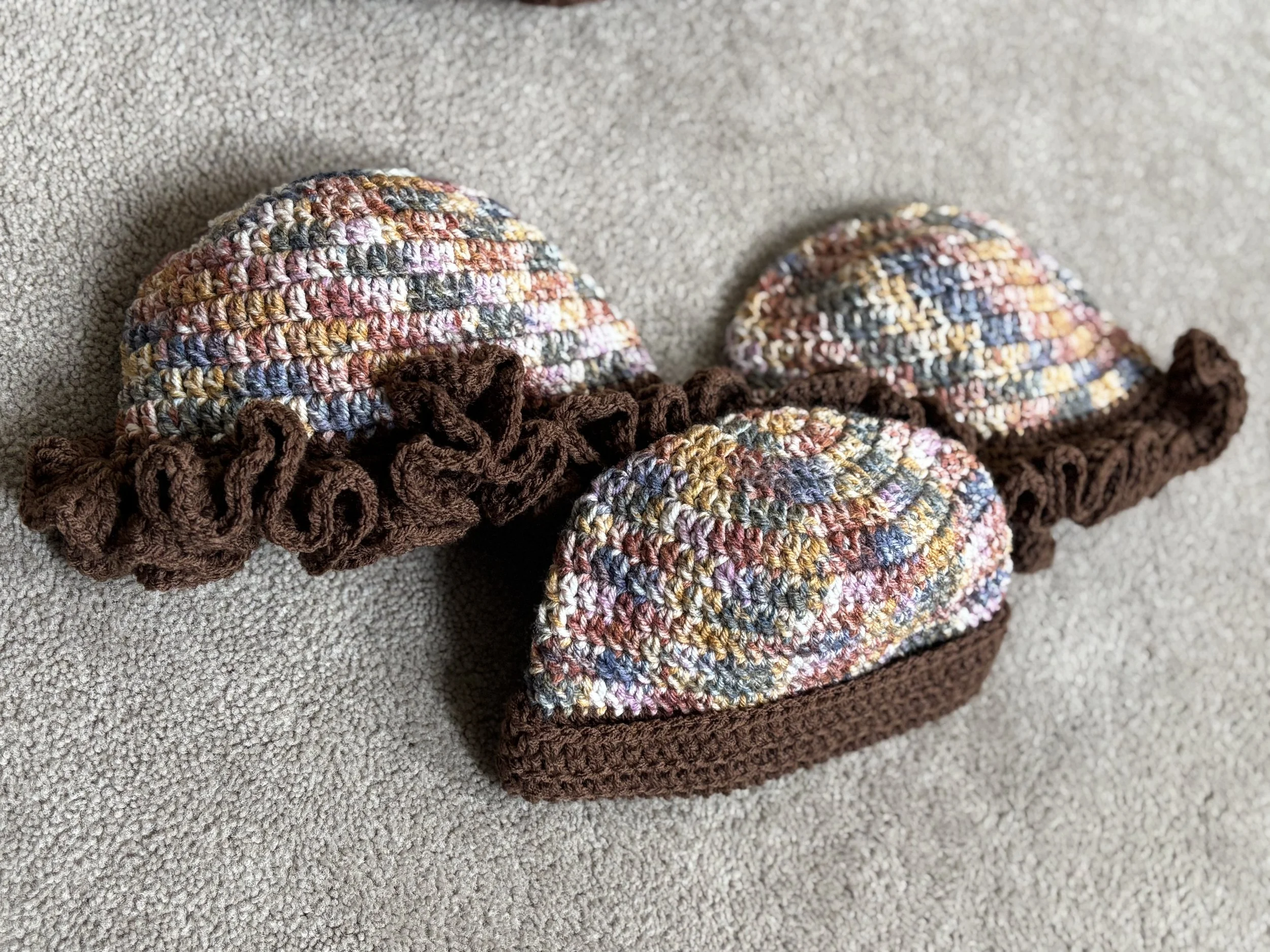 Three multicolored crocheted caps with ruffled brims displayed on a light gray carpet.