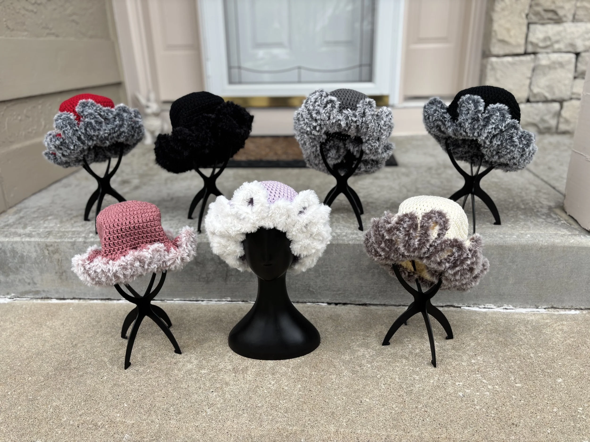 A collection of handmade crochet hats with fluffy edges displayed on black mannequin heads outside a house.