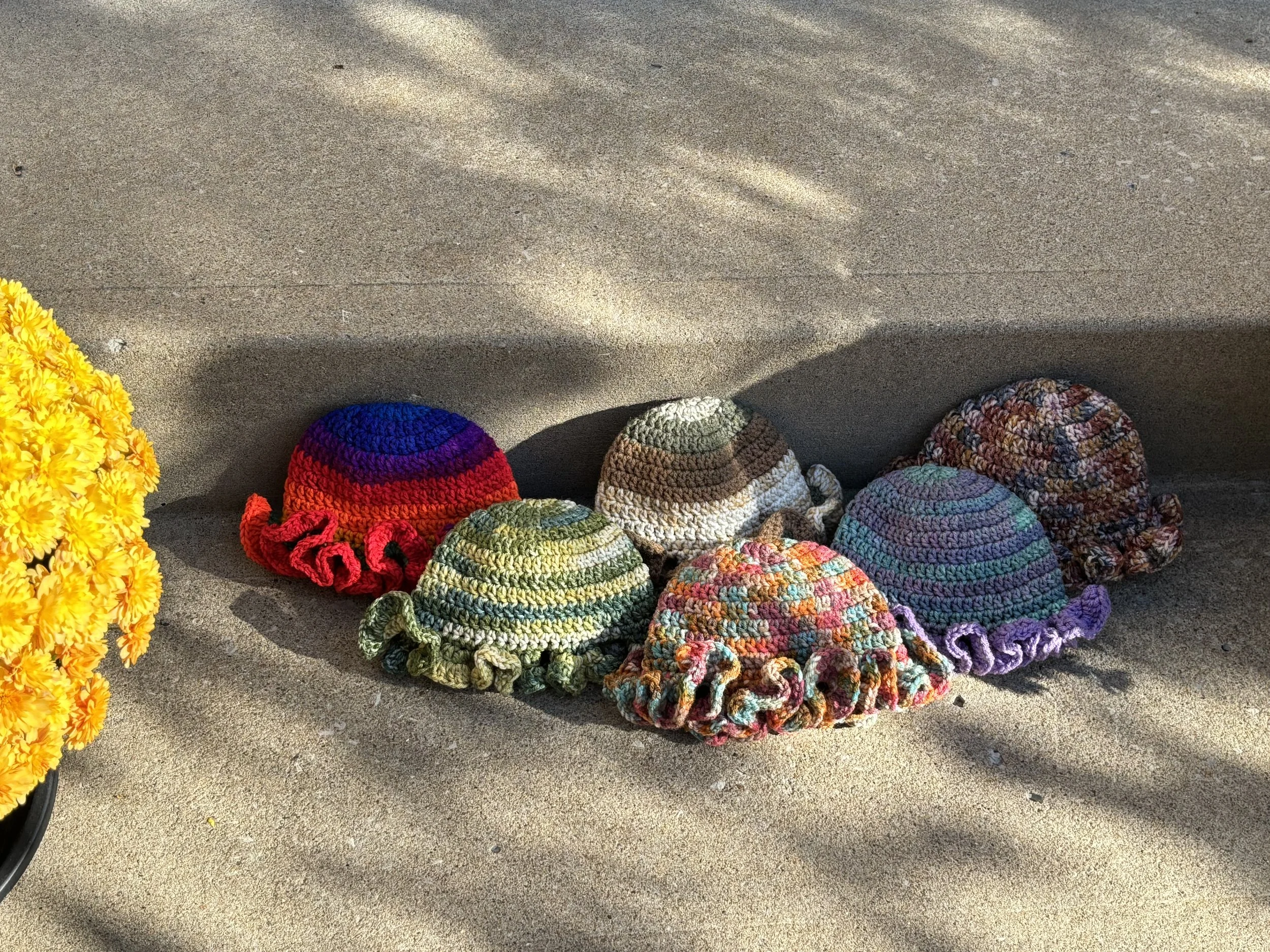 Five colorful crocheted hats with ruffled brims on a concrete step next to a pot of yellow flowers.