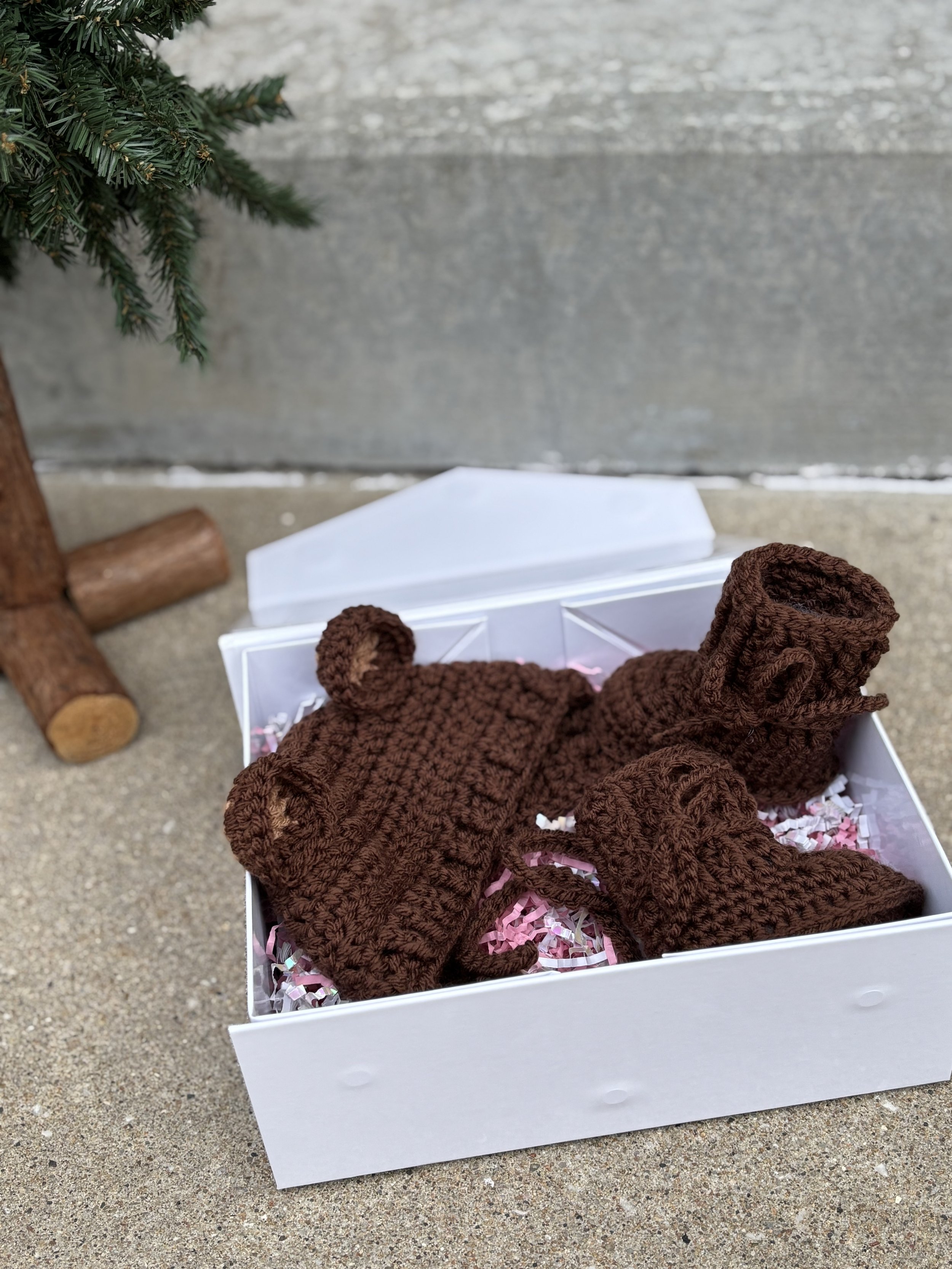 A gift box with handmade brown knitted baby items, including a hat with bear ears and booties, placed on a textured ground next to a wooden tree base.