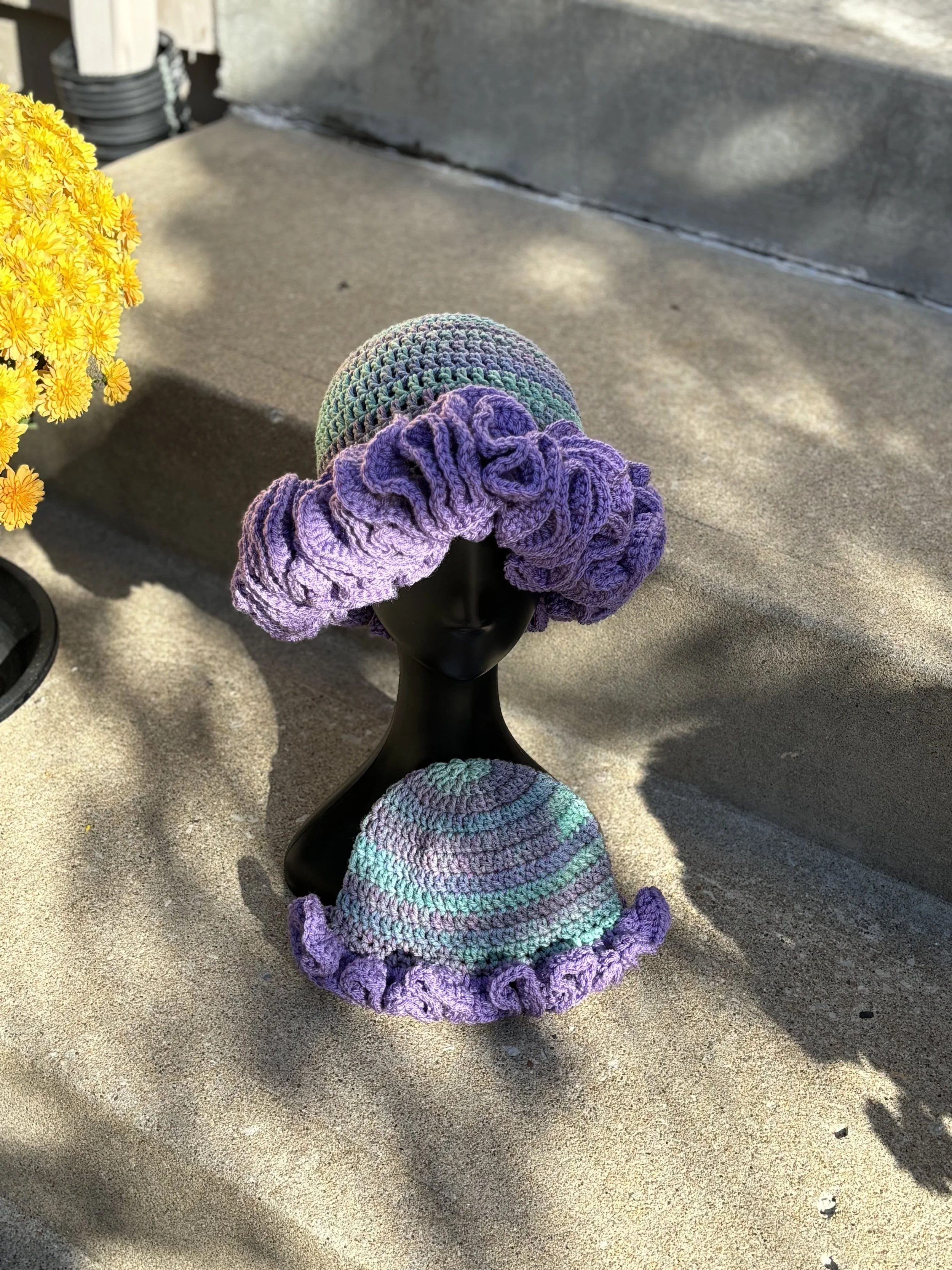 Crocheted hats on a mannequin head with purple ruffled edges and green and purple stripes, displayed on a concrete step next to yellow flowers.