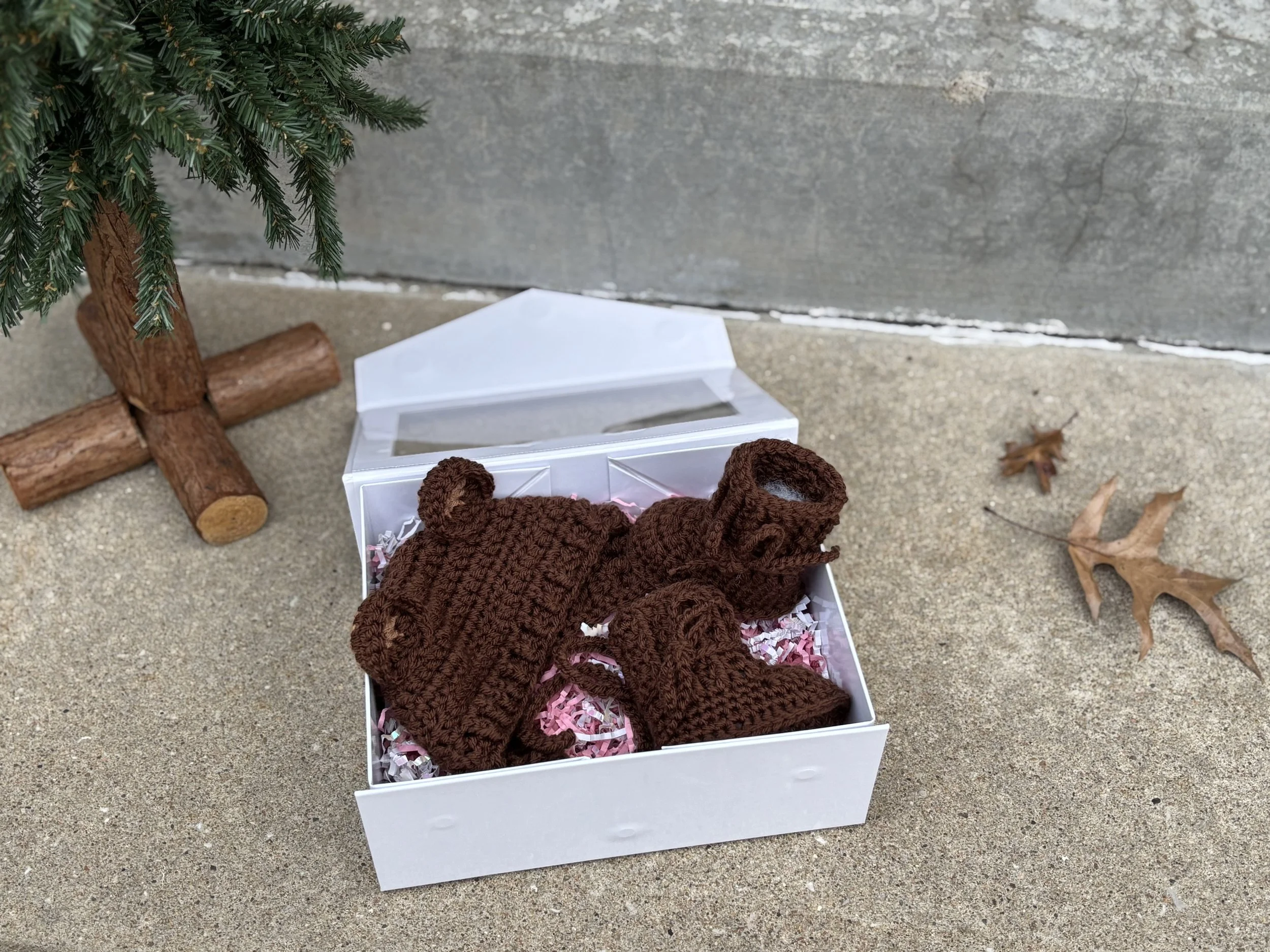 Open white gift box containing a crocheted brown hat with bear ears and matching booties, placed on a textured surface next to a small artificial Christmas tree and dry leaves.