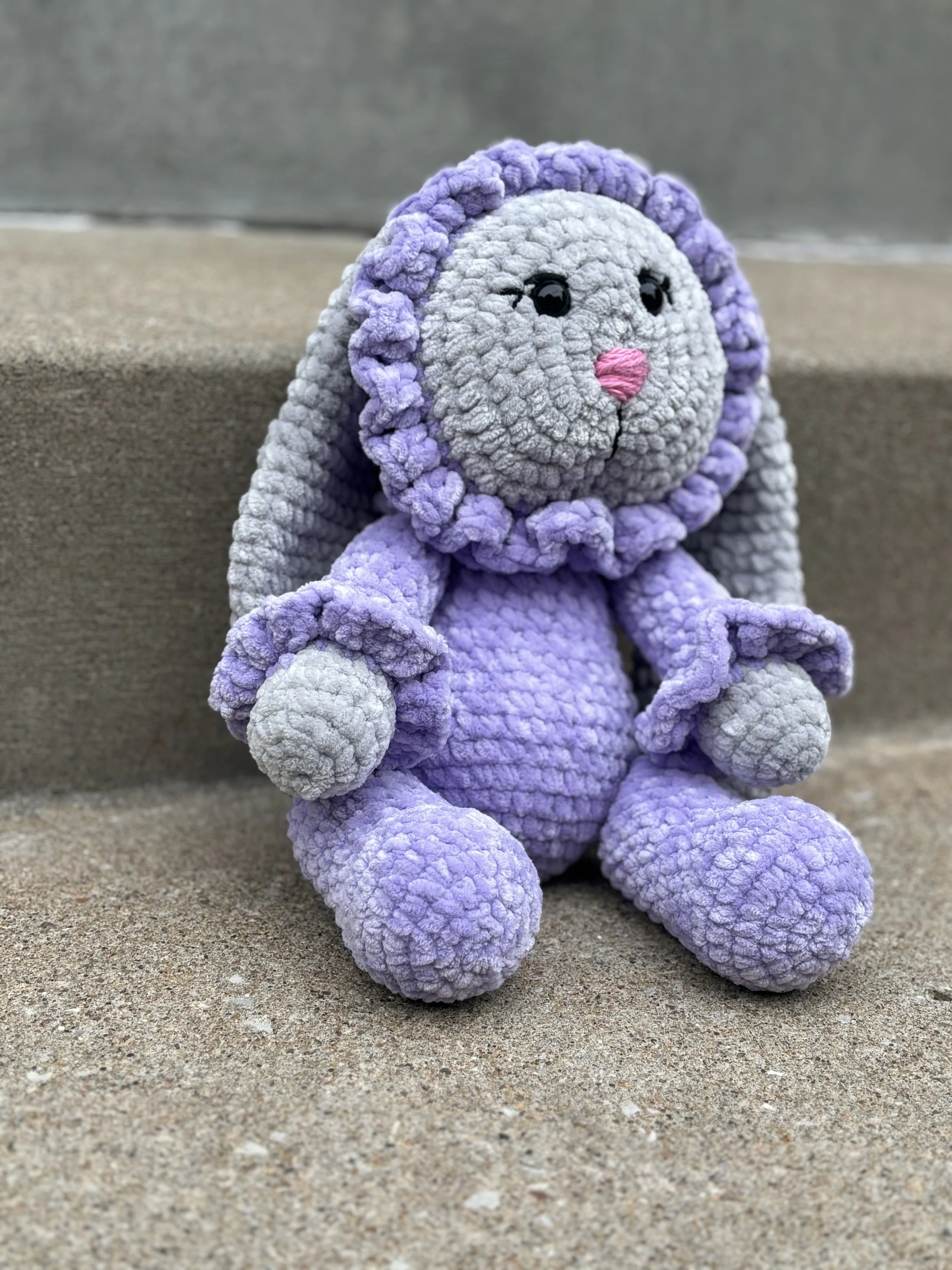 Crocheted stuffed bunny with purple and gray yarn sitting on concrete steps.