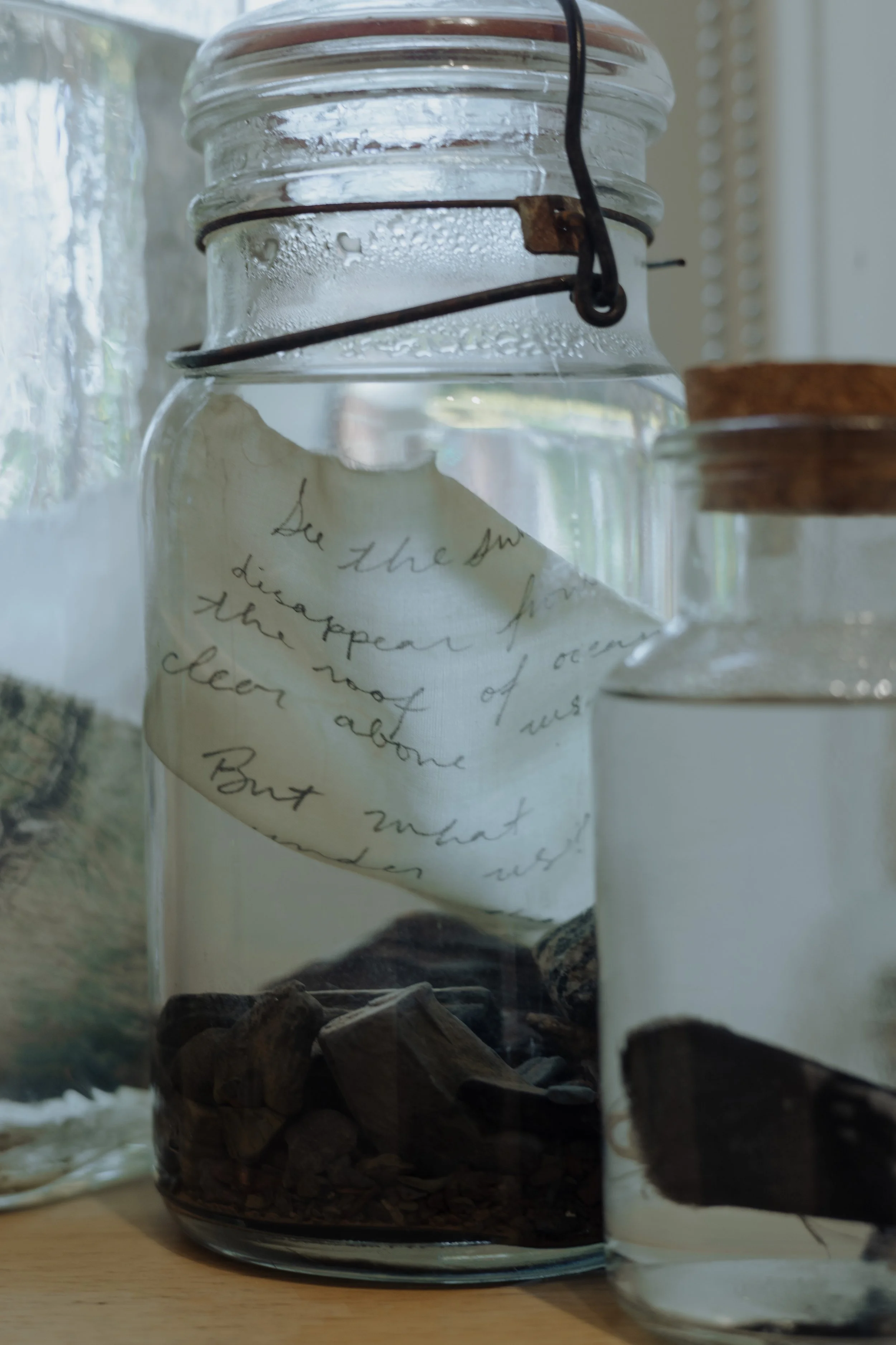 A glass jar with a handwritten note floating inside, surrounded by other jars with cork lids, all on a wooden surface.