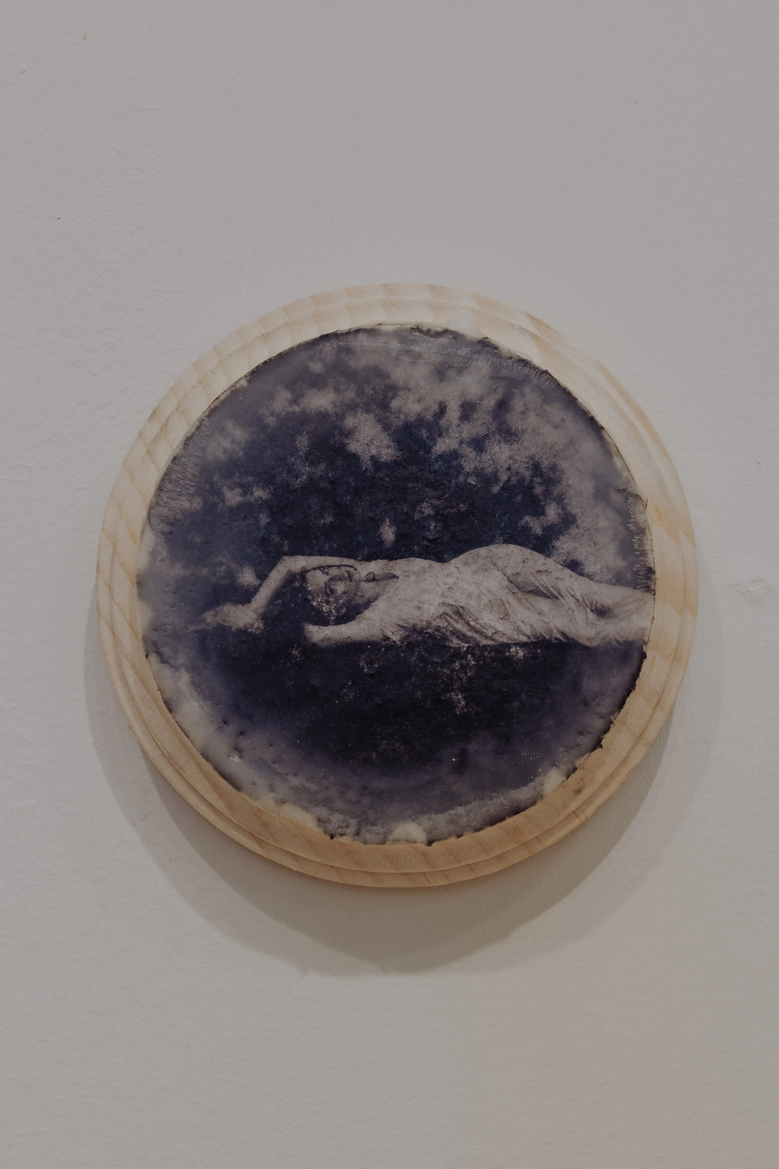Art piece on a round wooden frame depicting a reclining woman with clouds in the background.