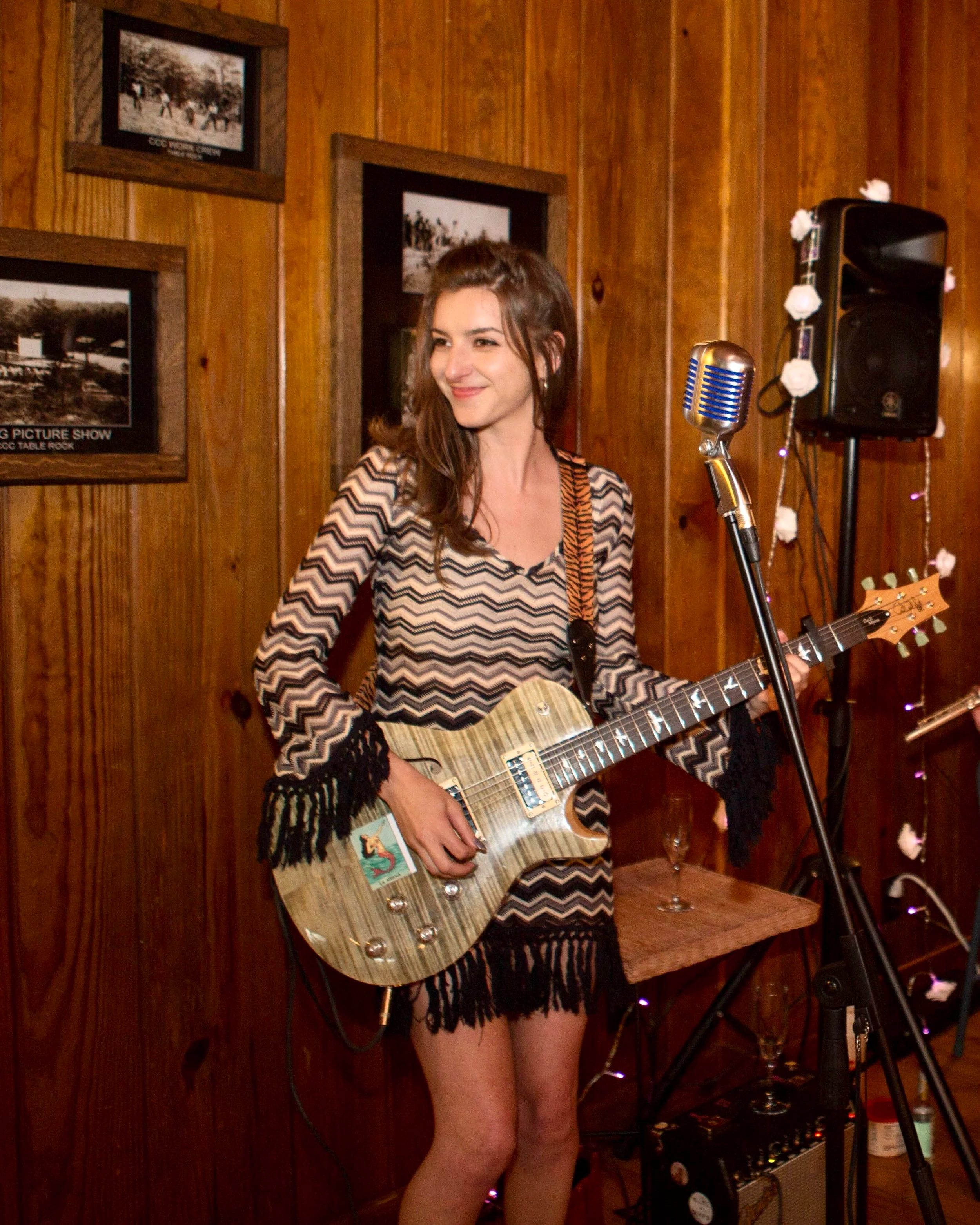 A young woman with long brown hair, smiling, playing an electric guitar in a wooden-paneled room decorated with framed black and white photos, a microphone stand, a speaker, and fairy light decorations.
