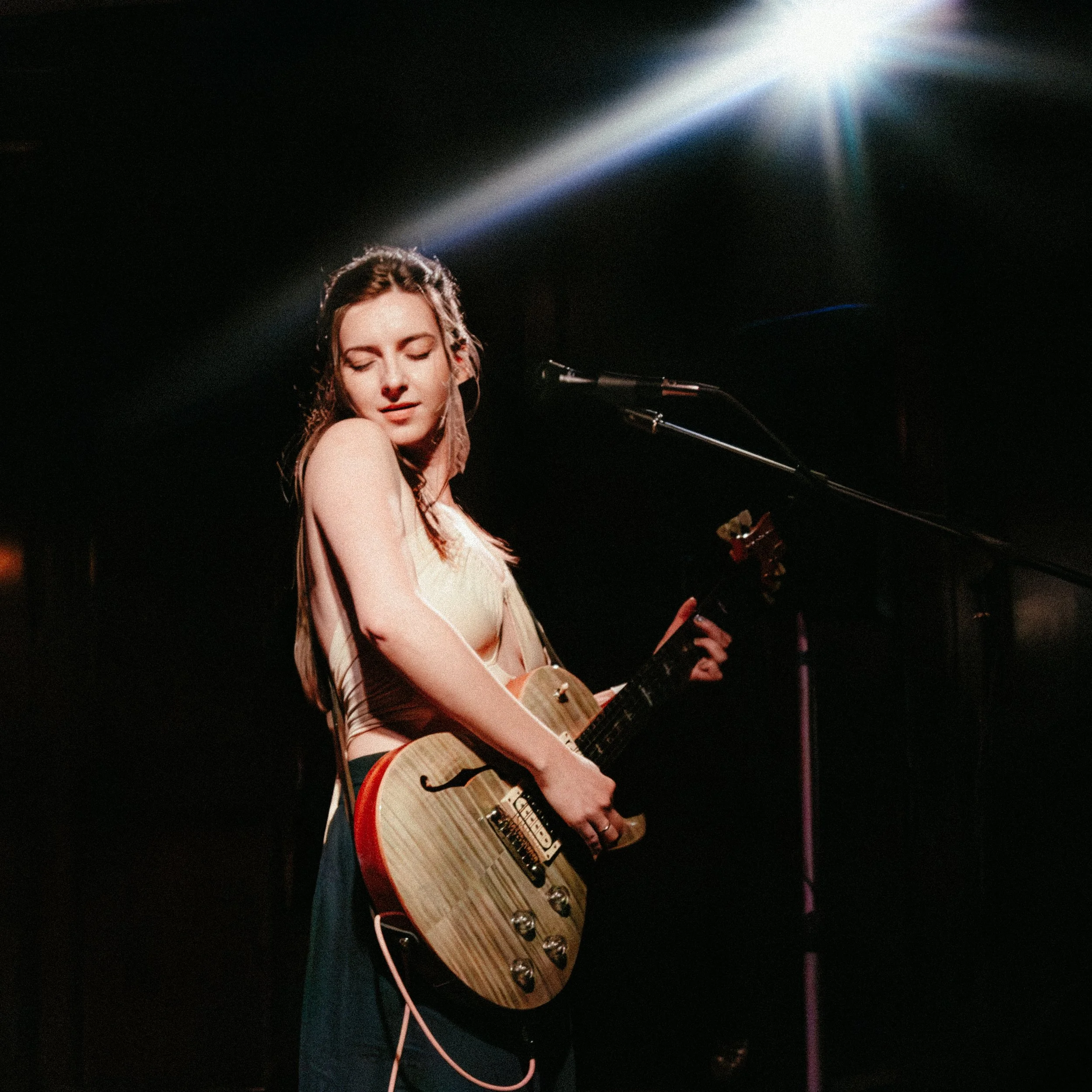 A young woman with long hair, wearing a sleeveless top and dress, playing an electric guitar on stage with a spotlight shining down from above.