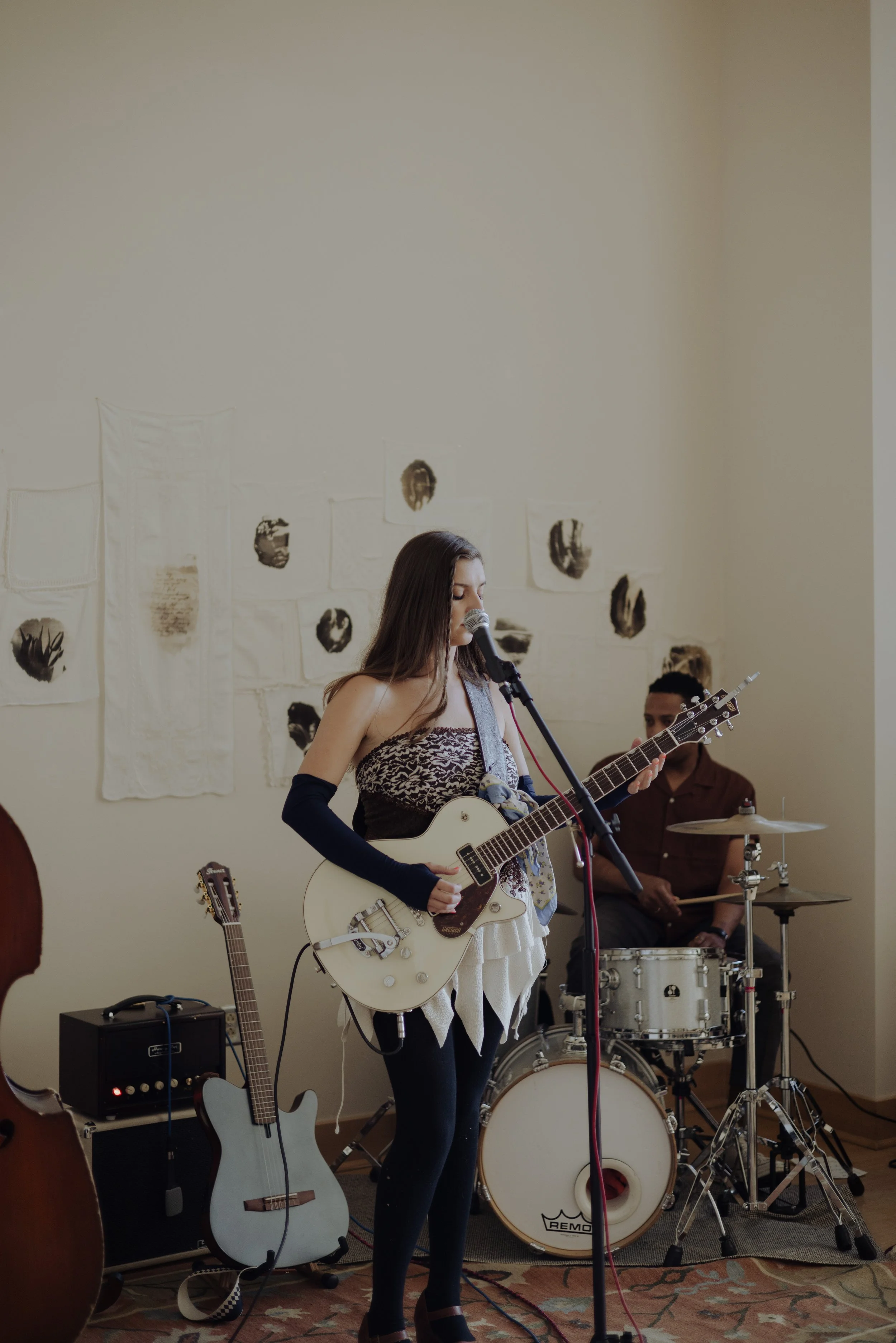 A young woman is playing an electric guitar and singing into a microphone during a live music performance, with a man playing drums in the background and artwork on the wall behind them.