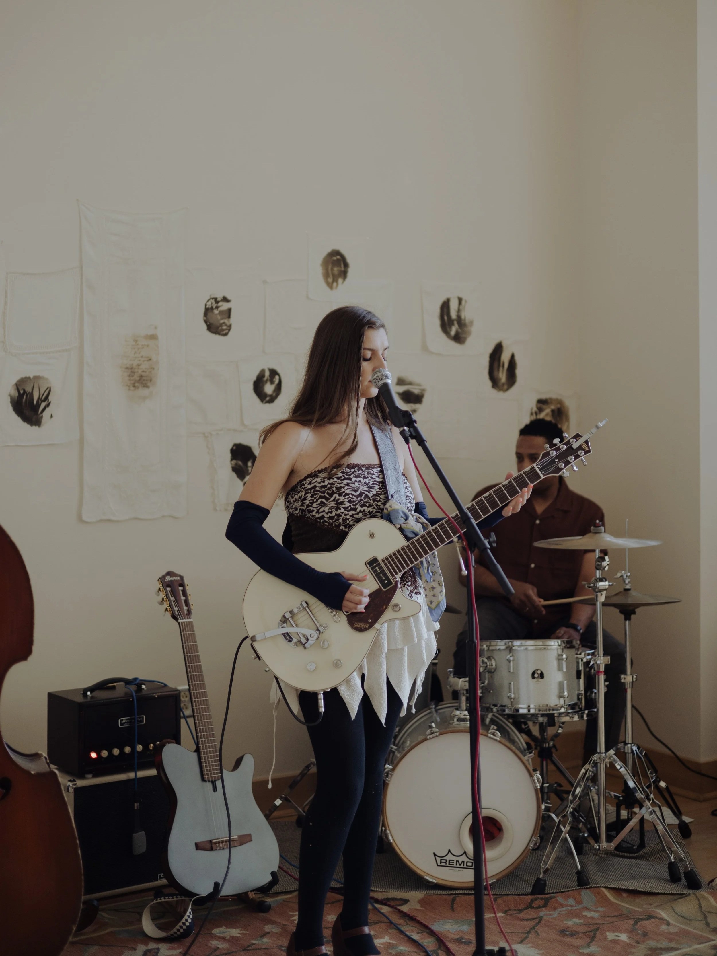 Young woman playing an electric guitar and singing into a microphone, with a drummer in the background, in an indoor performance setting.