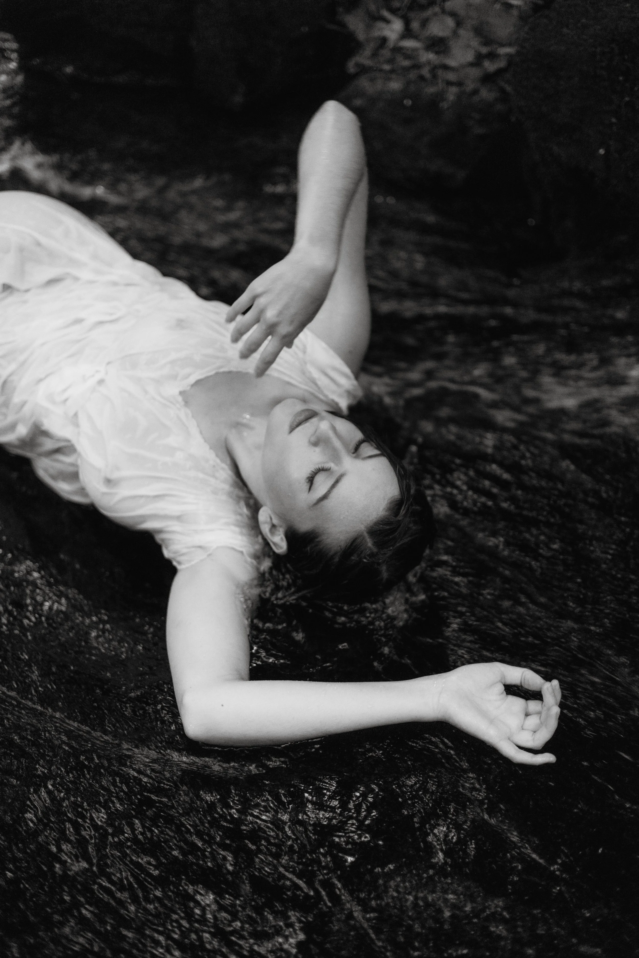 A woman with short dark hair and light skin fragment, wearing a light-colored dress, lies in shallow water with her eyes closed. Her arms are extended with one hand resting above her head and the other gently touching her face, creating a serene and tranquil scene in black and white.