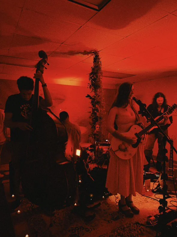 Band performing live music on a stage with red lighting, featuring a woman playing guitar and singing, and a man playing upright bass with a doll on top, in a cozy indoor setting.