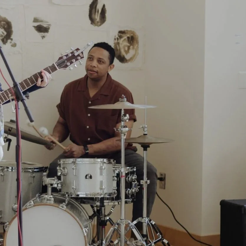 A man sitting behind a drum set holding a stick, with a person playing an acoustic guitar nearby, in a room with abstract wall art.