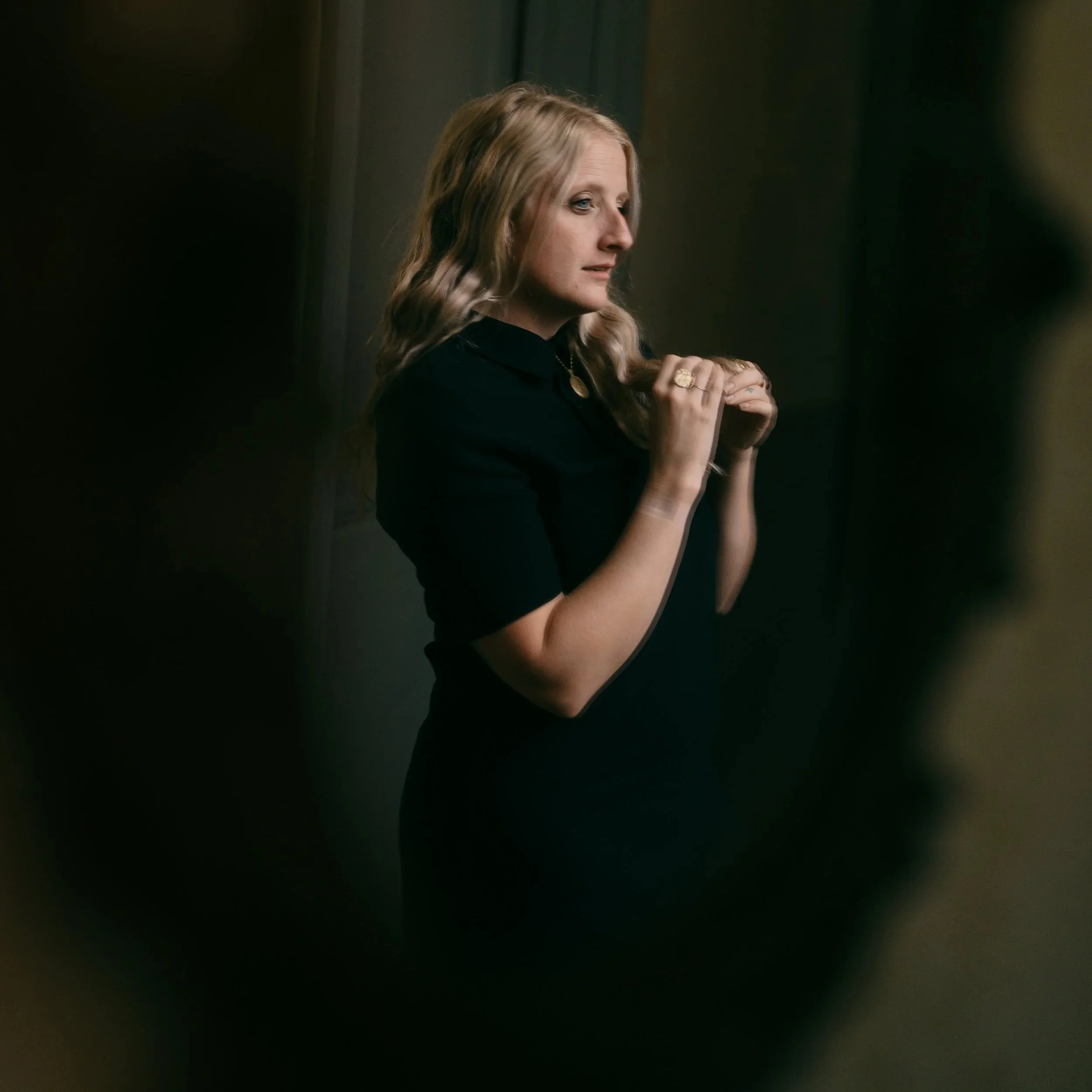 A woman with long, wavy blonde hair wearing a black dress, standing with her hands clasped near her collar, in a dark room with blurred dark shapes in the foreground.
