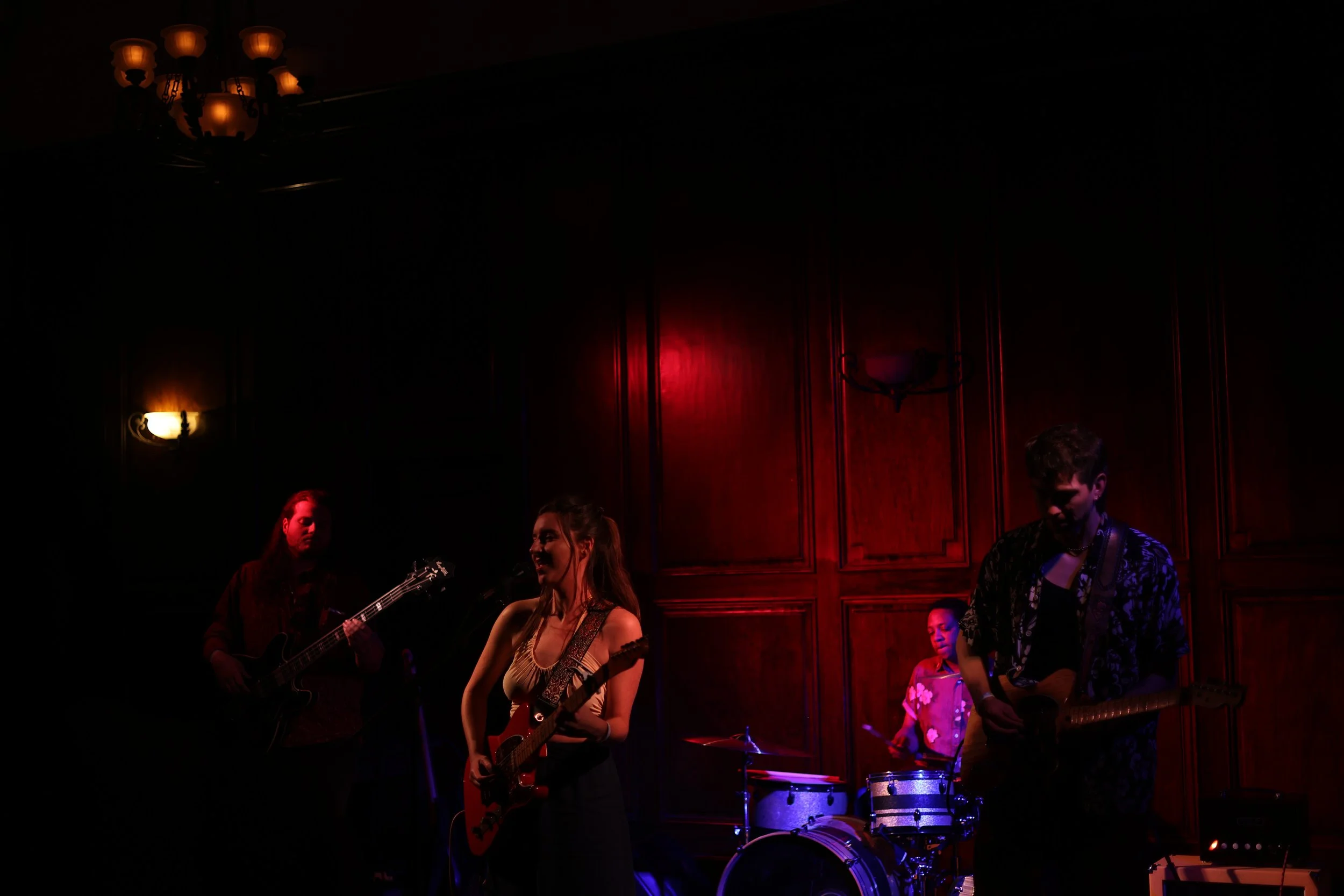 Group of four musicians performing on stage, with a dark wood-paneled background, including two guitarists, a drummer, and a bassist, illuminated by warm stage lighting.