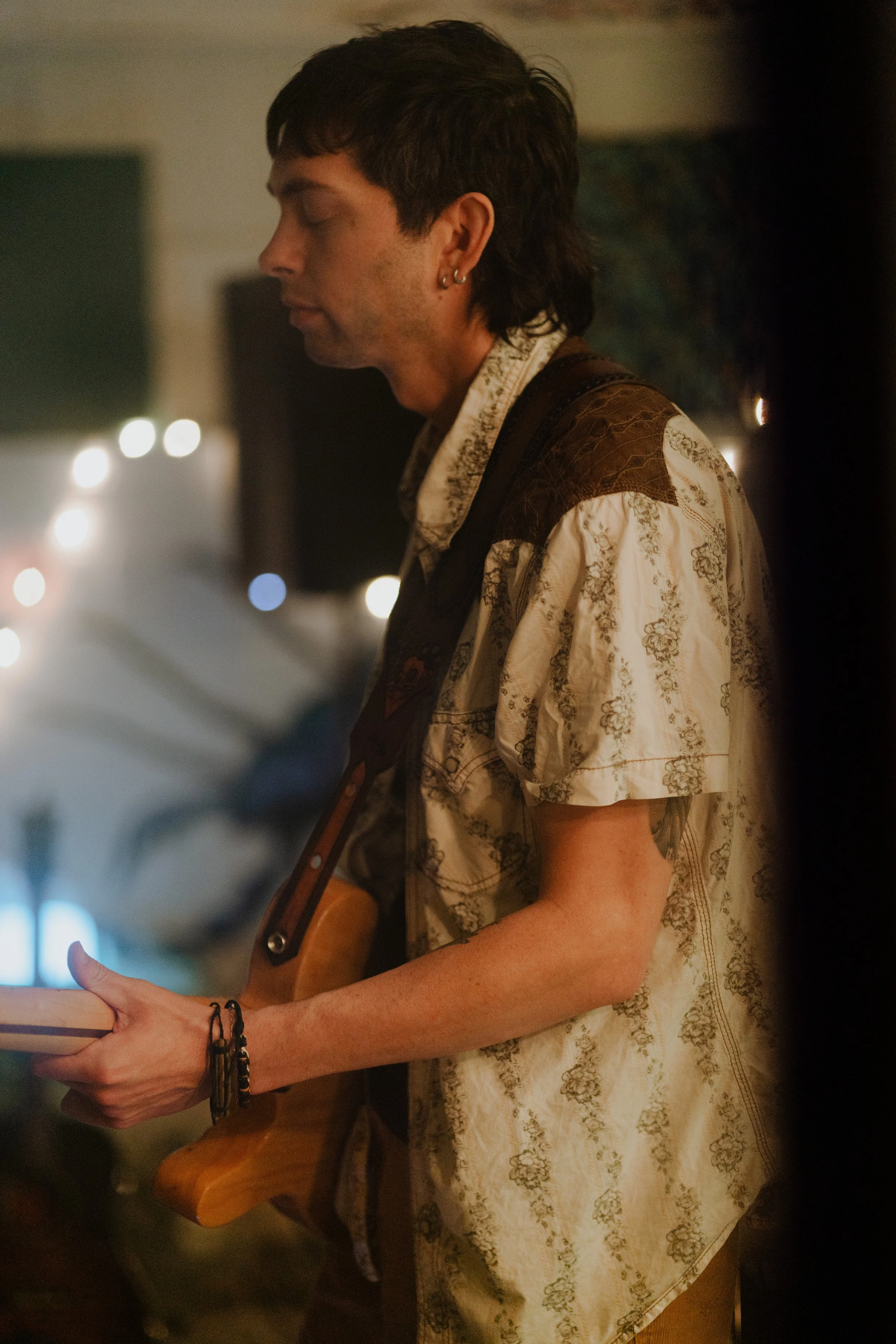 A man with dark hair and earrings plays an electric guitar, eyes closed, in a dimly lit, cozy room with blurred background lights.