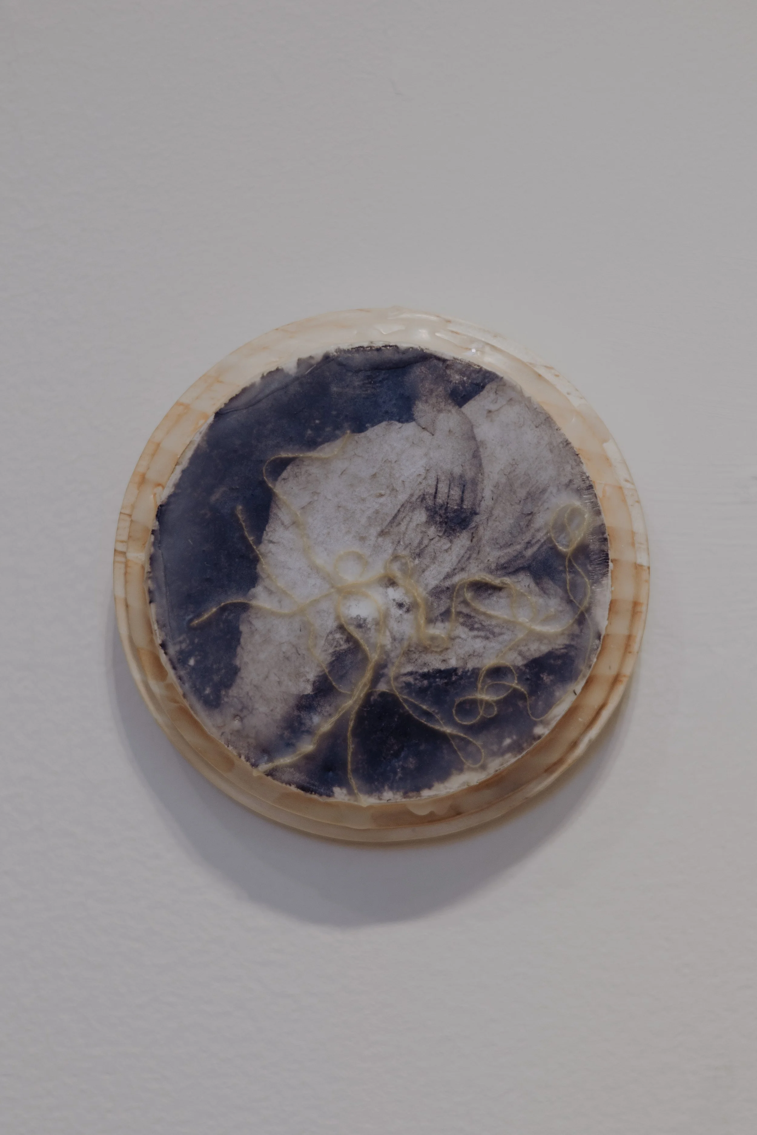 A circular object with black, white, and cream-colored marbled patterns, embedded with thin, tangled gold-colored threads, mounted on a white wall.