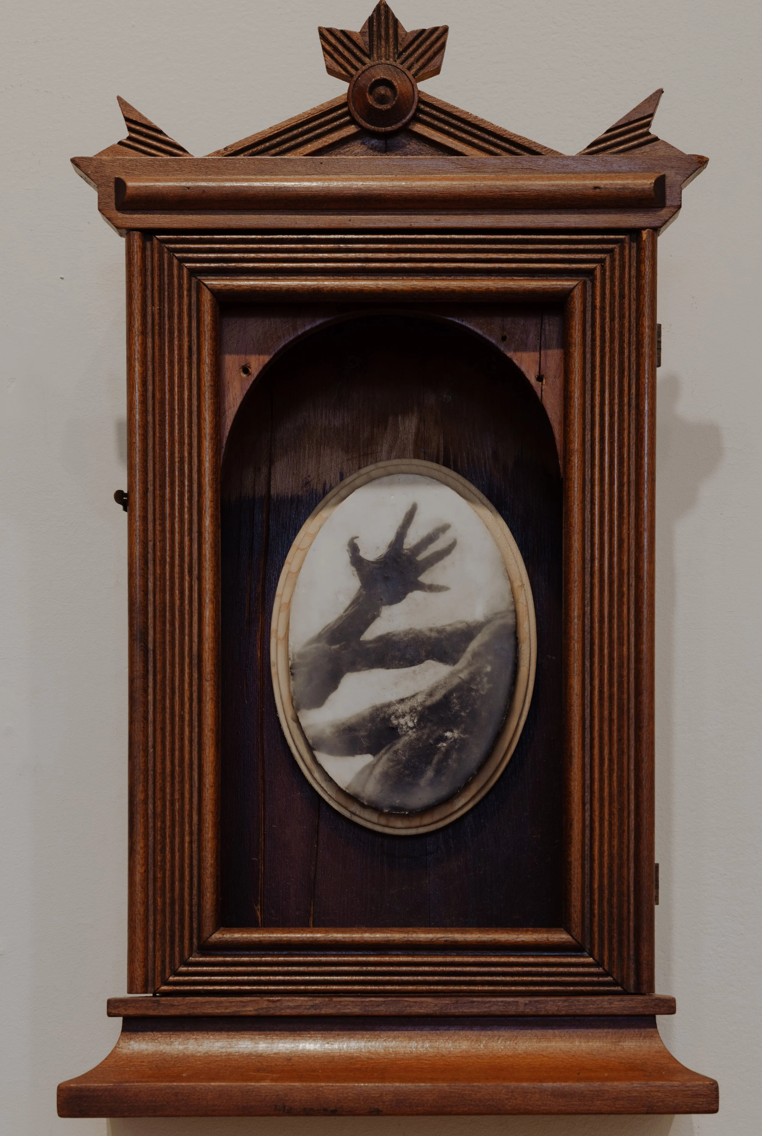 A vintage wooden wall clock with a dark silhouette of a hand reaching out, mounted on a light-colored wall.