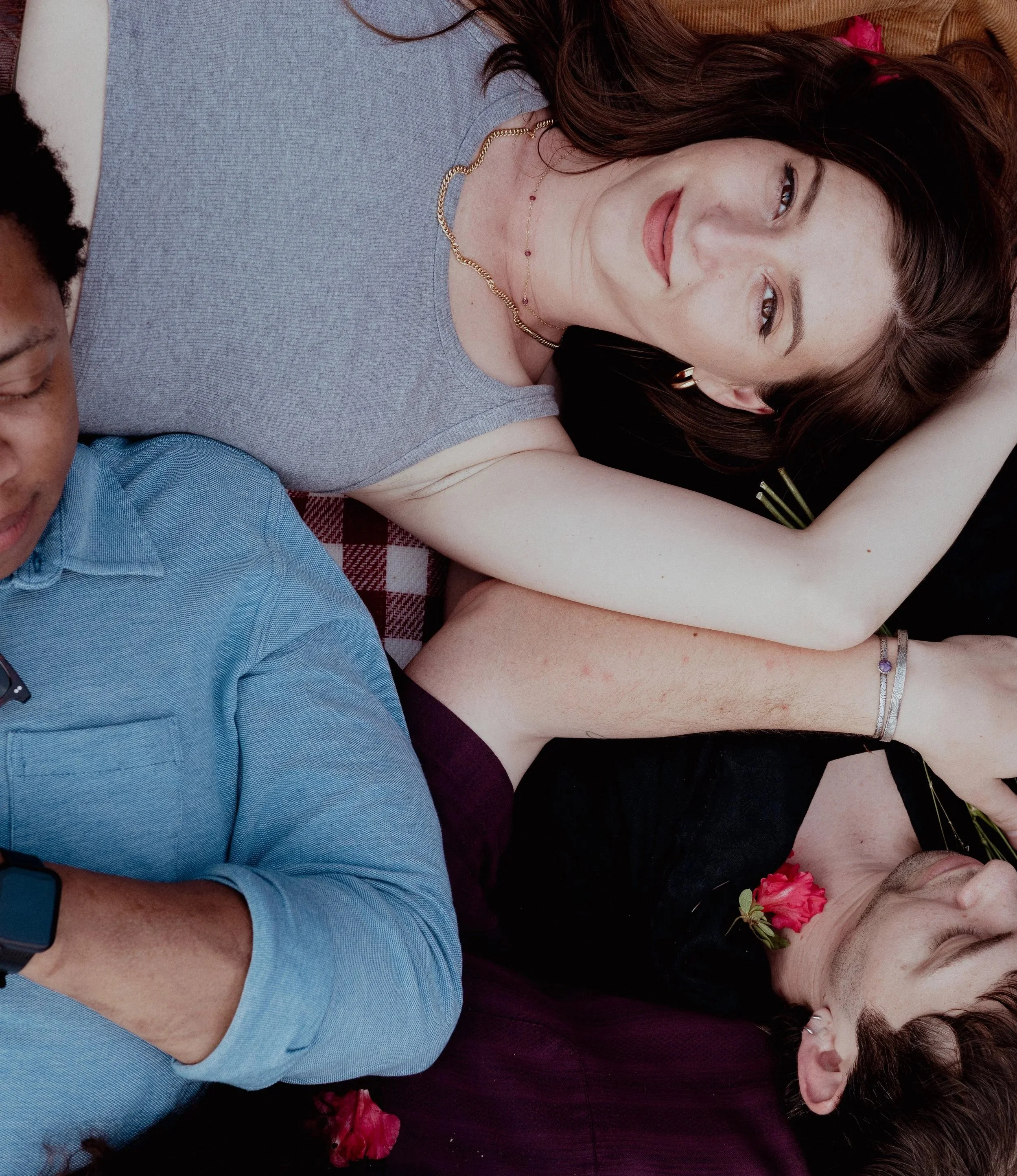 Three young adults lying on the ground close together, with one woman in the center smiling and resting her head on her arm, and two men beside her, one with a flower near his chest.