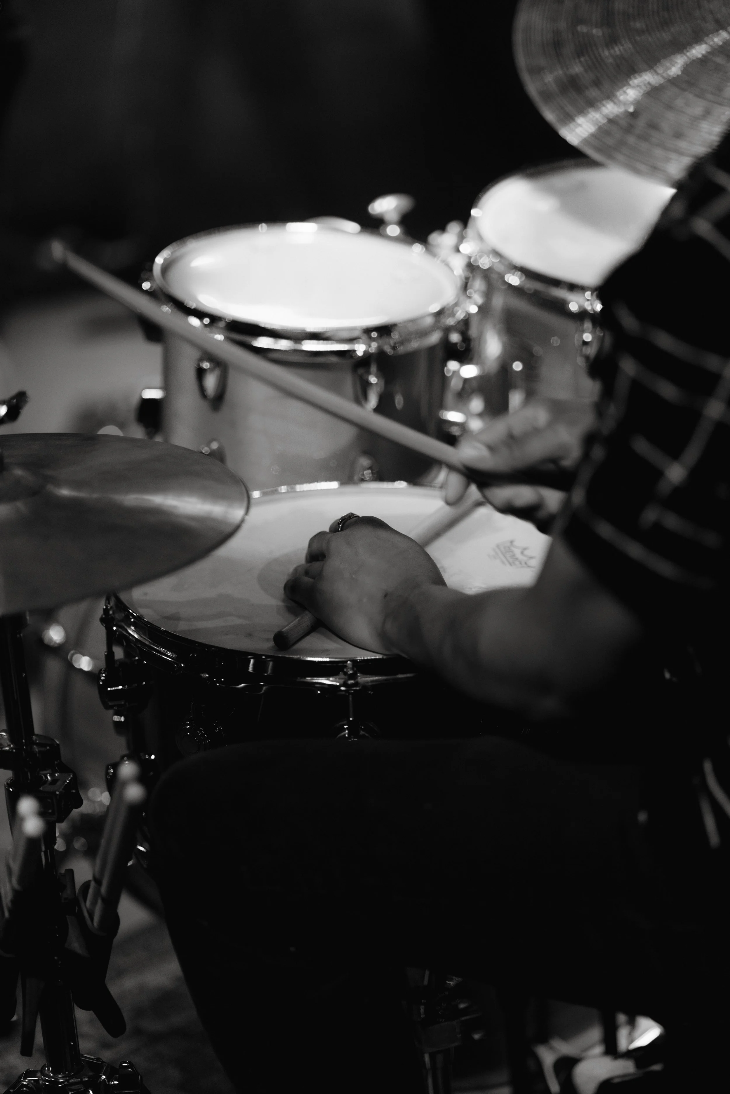 A person playing the drums, holding a drumstick in their hand, with a drum kit in the background. The image is in black and white.