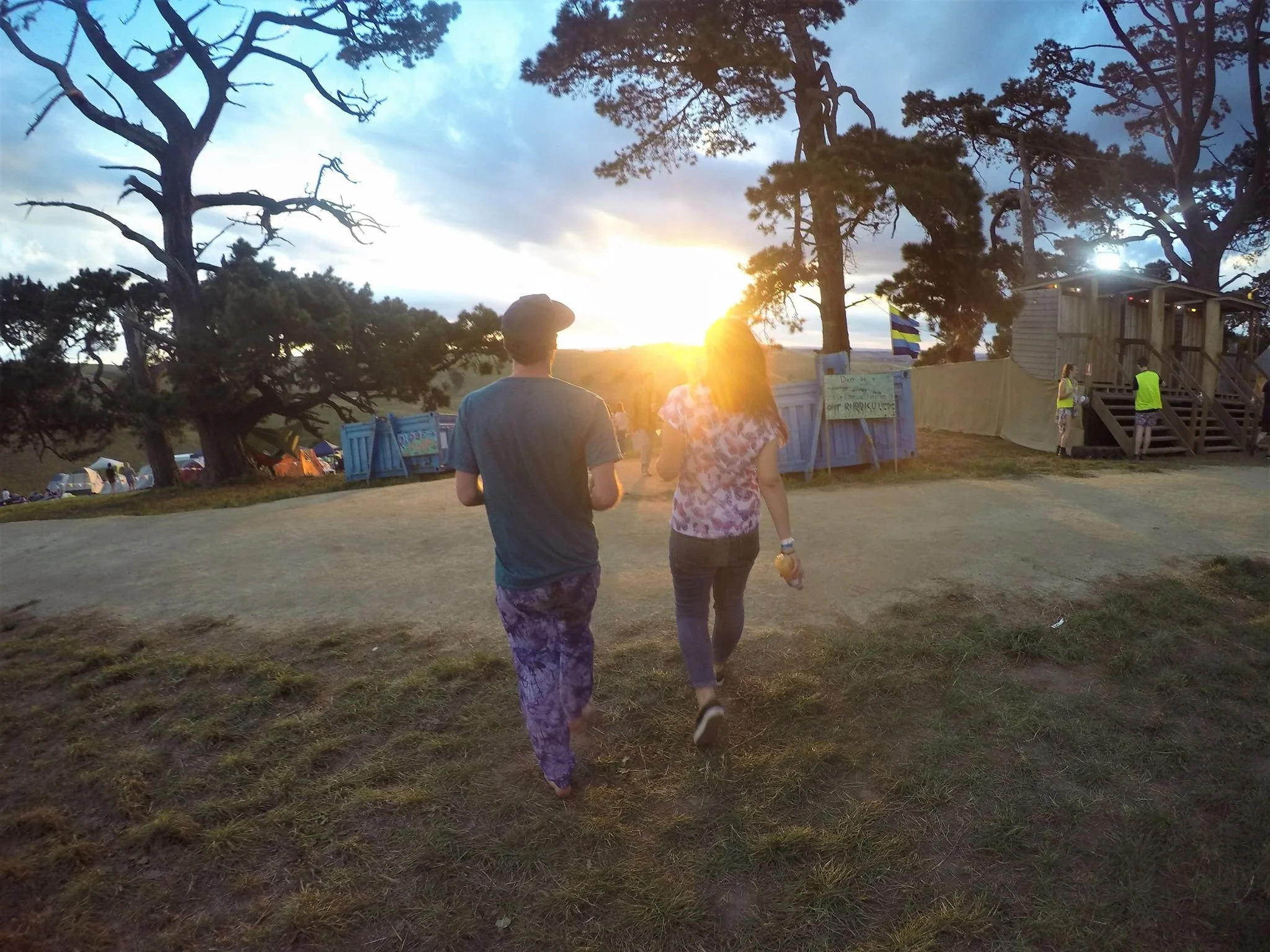 Two people walk away from the camera on a grassy path toward a sunset scene with trees, tents, and a wooden structure in the background.