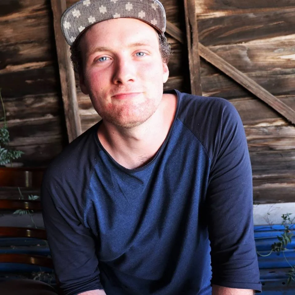 Young man with light skin and blue eyes wearing a gray cap and a navy and black long-sleeve shirt, smiling slightly, sitting in front of a wooden background.