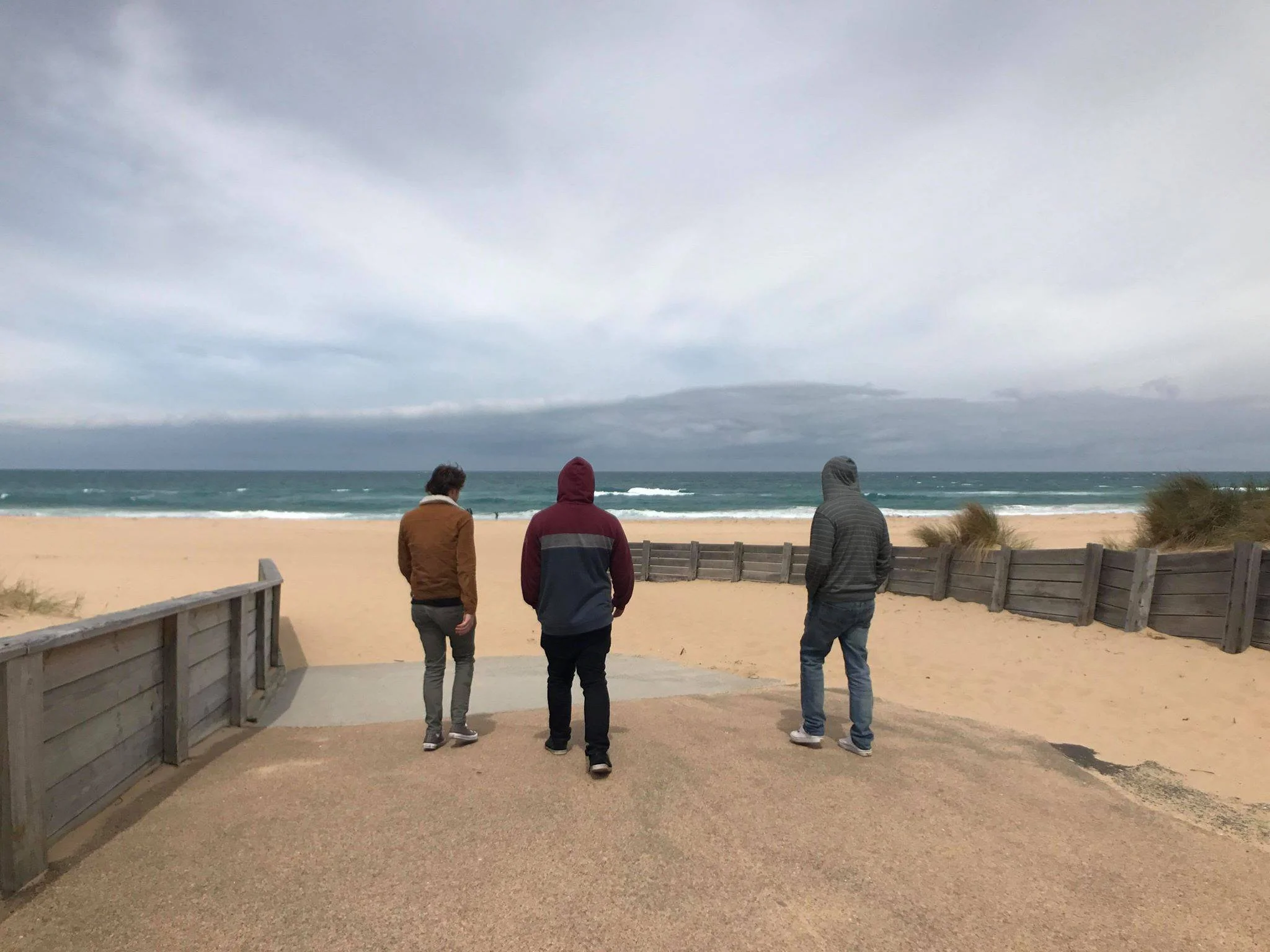 Three people in hooded jackets standing on a pathway facing the beach and ocean on a cloudy day, with sand dunes and a wooden fence on either side.