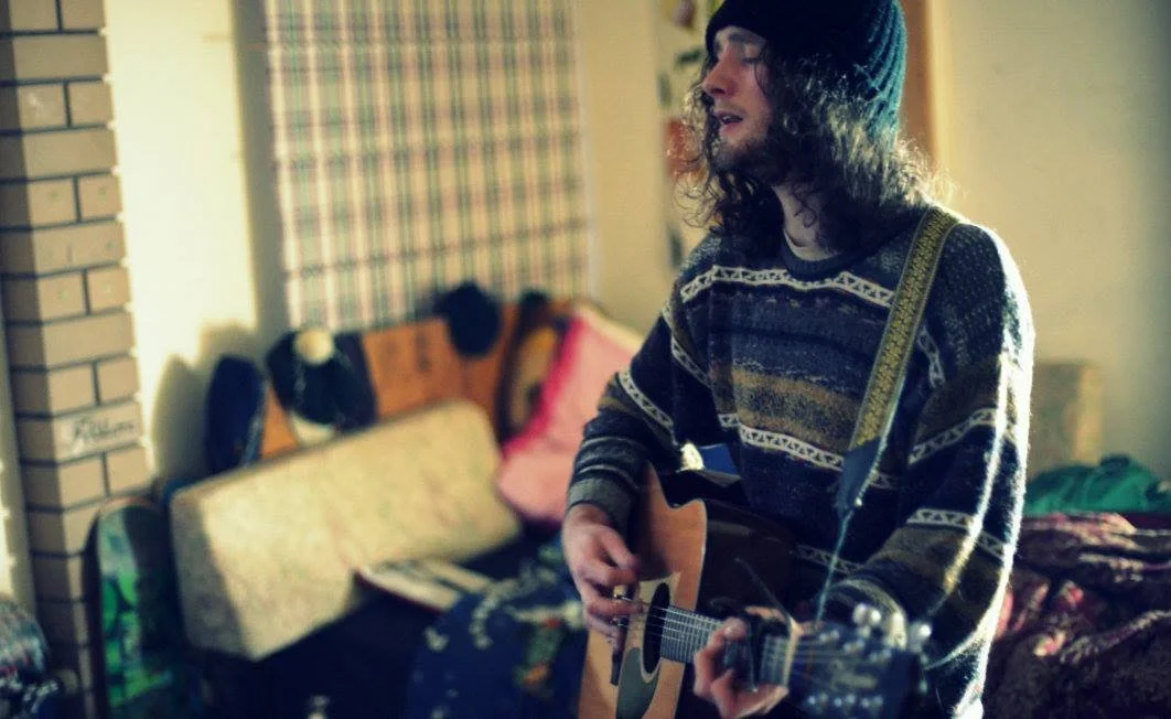 A young man with long curly hair, sunglasses, and a beanie playing an acoustic guitar indoors.