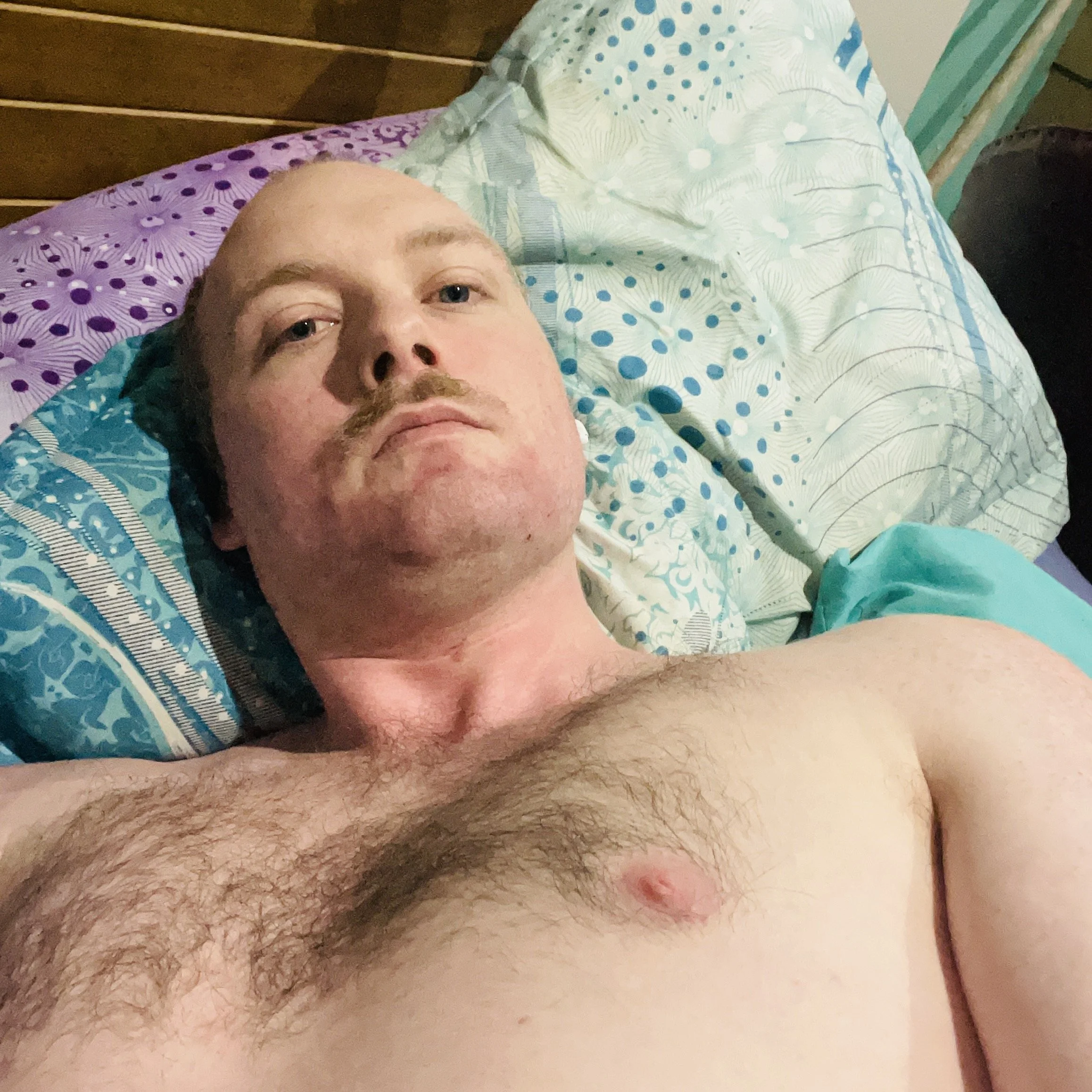 A topless man with light skin and a mustache lying on a bed, looking at the camera.