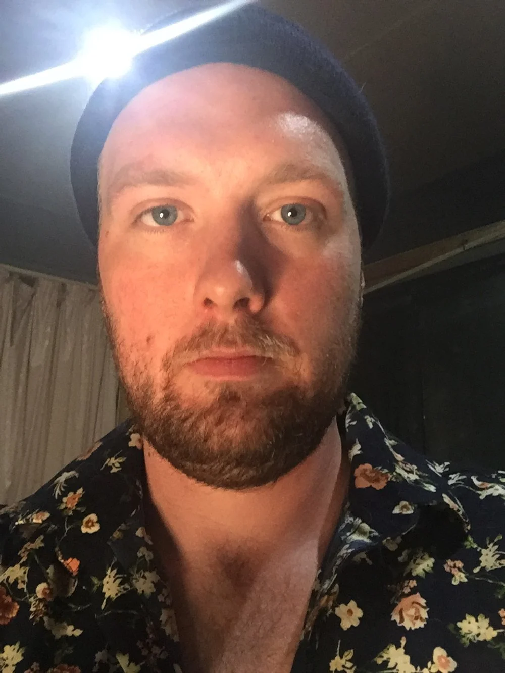 Close-up of a man with a beard and mustache, wearing a black beanie and a floral-patterned shirt, with neutral expression, indoors with dim lighting.