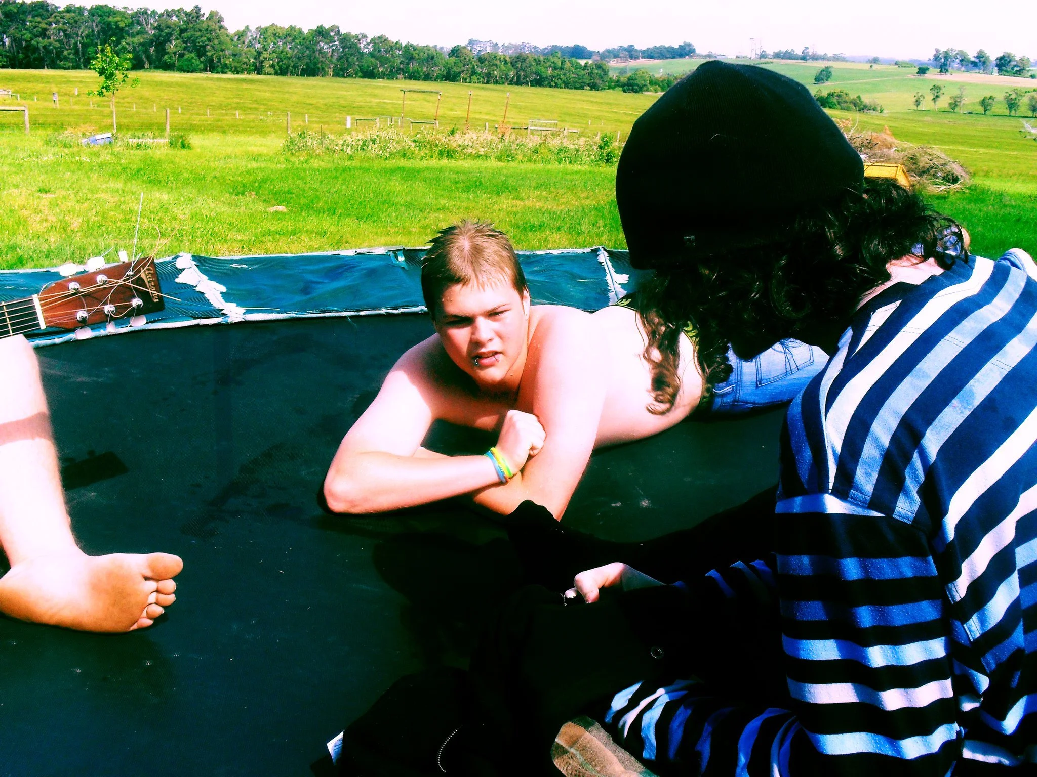 Young man lying on a trampoline outdoors with a landscape of green grass and rolling hills in the background, wearing only shorts, with a person in a striped shirt and a beanie sitting nearby.
