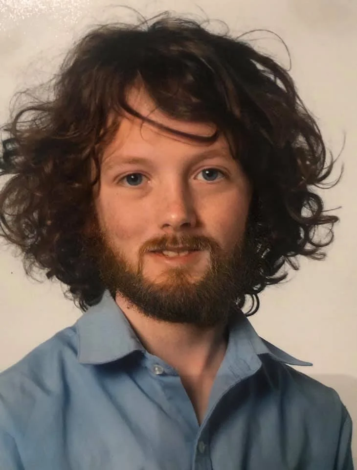 A man with curly brown hair, a beard, and blue eyes wearing a light blue collared shirt, standing against a plain background.