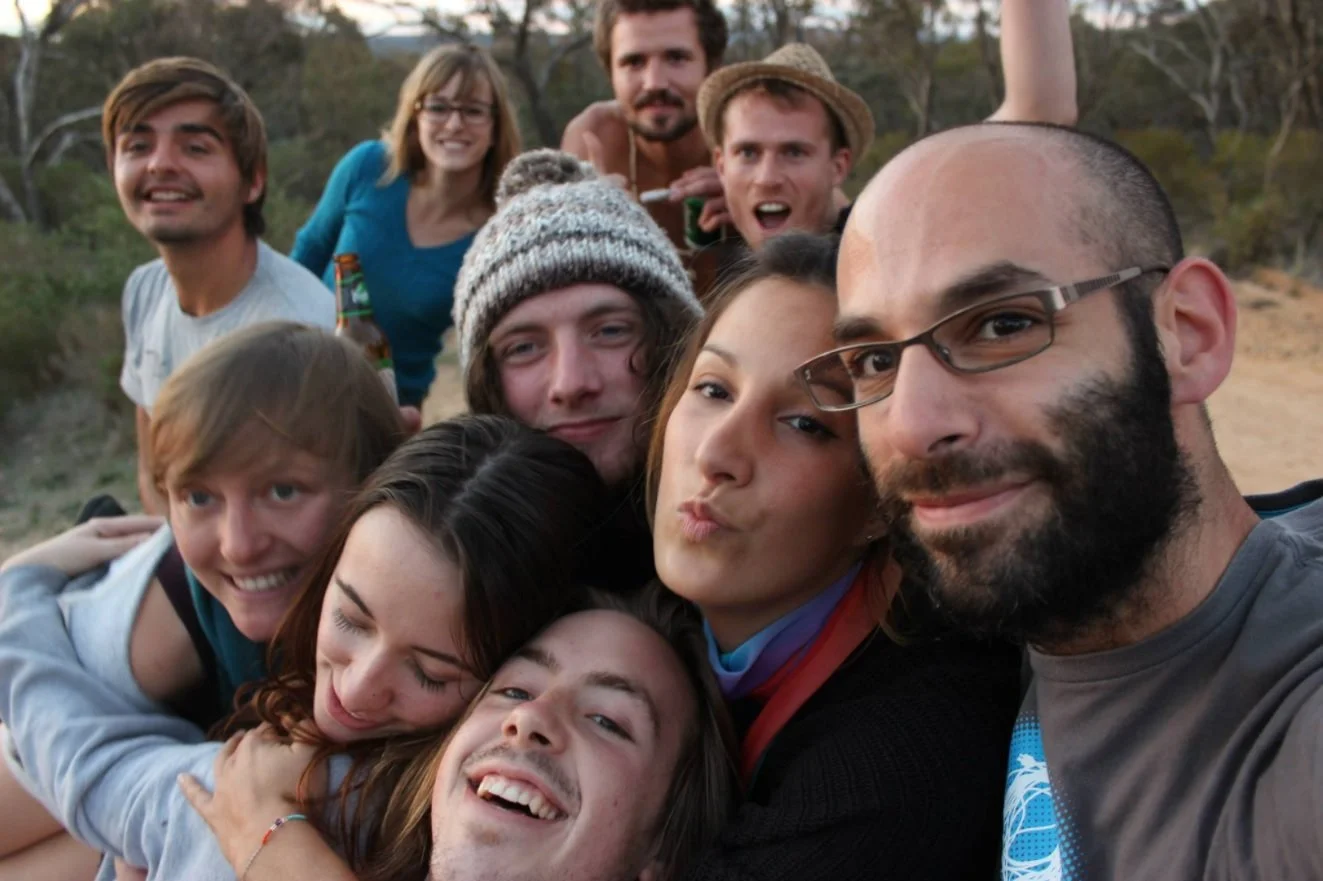 A group of people outdoors taking a selfie together in a natural setting with trees and a dirt path, smiling and making various facial expressions.