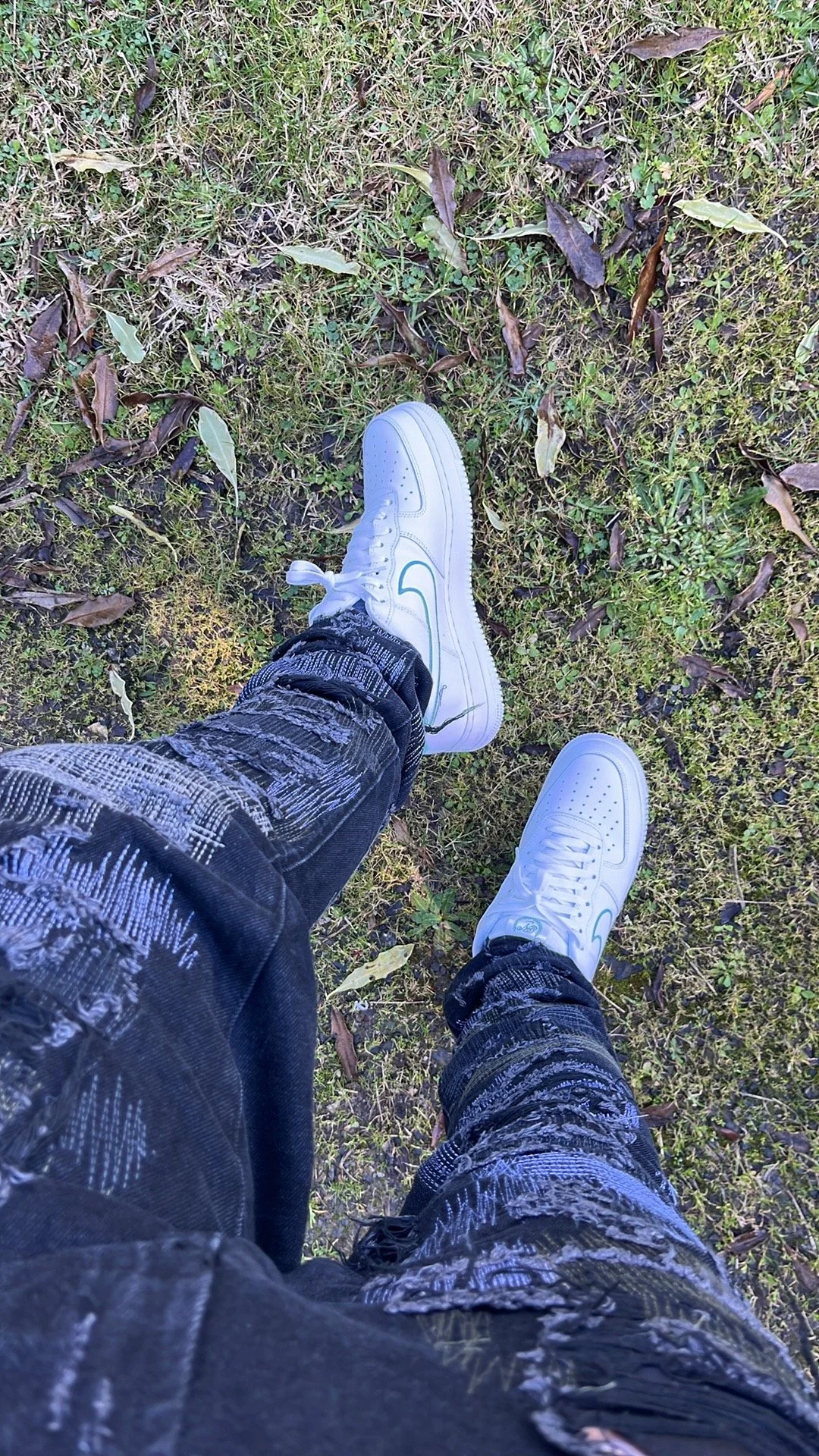 A person standing on grass and fallen leaves, wearing white Nike sneakers and ripped black jeans.