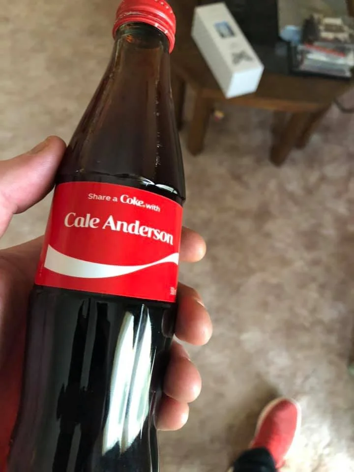 A person holding a small glass Coca-Cola bottle with a personalized label reading 'Share a Coke with Cale Anderson'; in the background, a wooden table, a white box, and some books are visible, with a beige carpeted floor and a red sneaker.