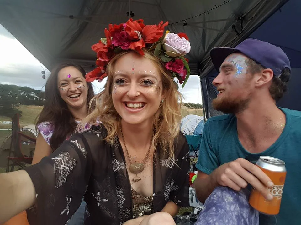 Three friends smiling at an outdoor gathering, with the woman in the center wearing a floral headpiece. They have glitter and face paint, and one is holding a drink can.