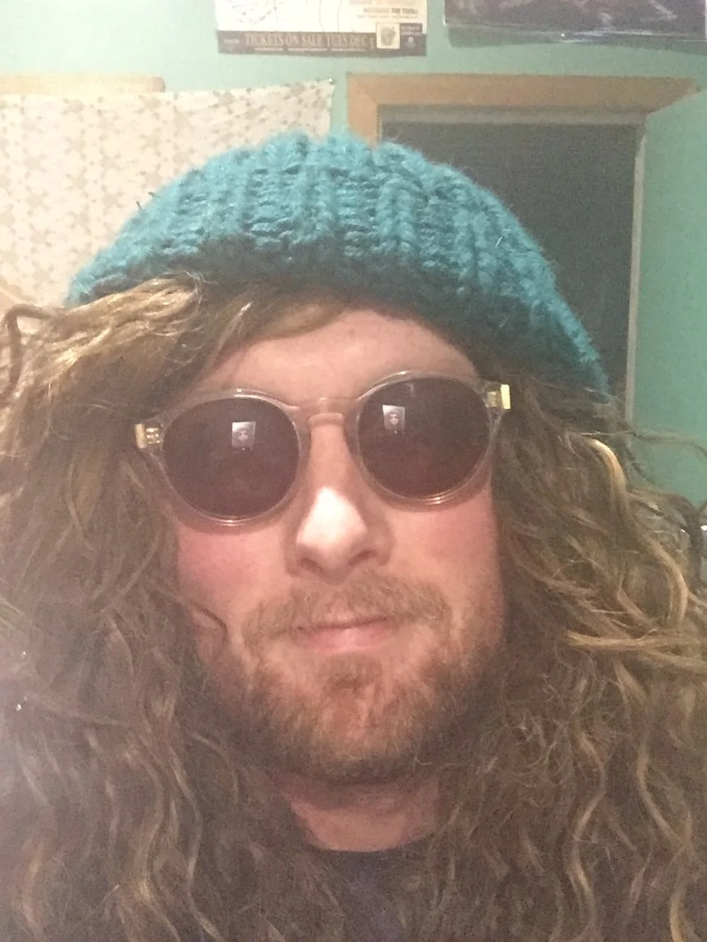 A person with long curly hair wearing round sunglasses and a blue knit beanie inside a room.