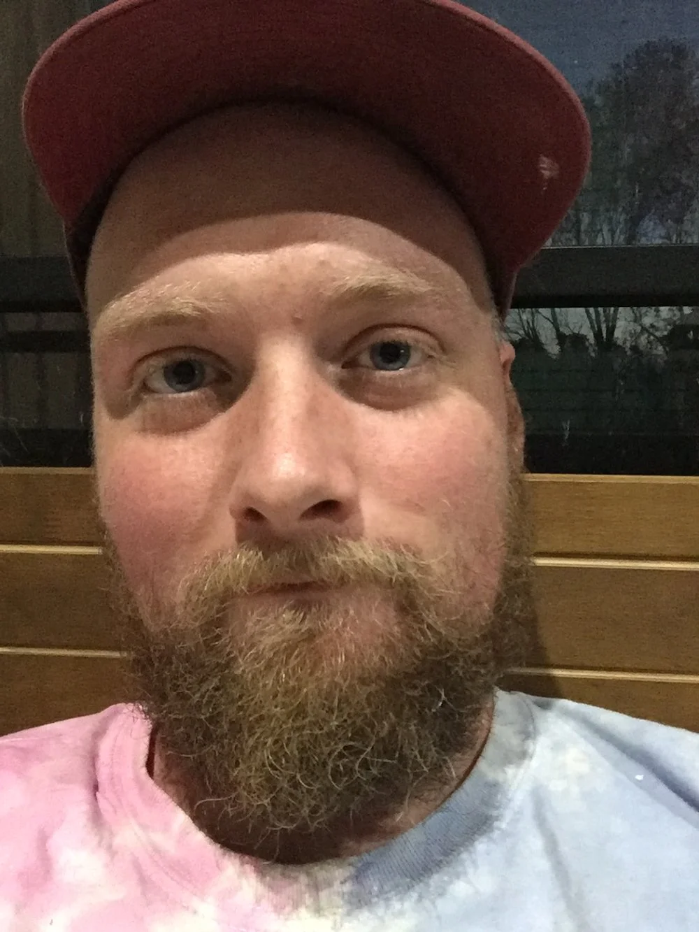 A man with a red cap, light blue eyes, and a full beard and mustache, sitting indoors with a wooden wall behind him and a window showing trees outside.