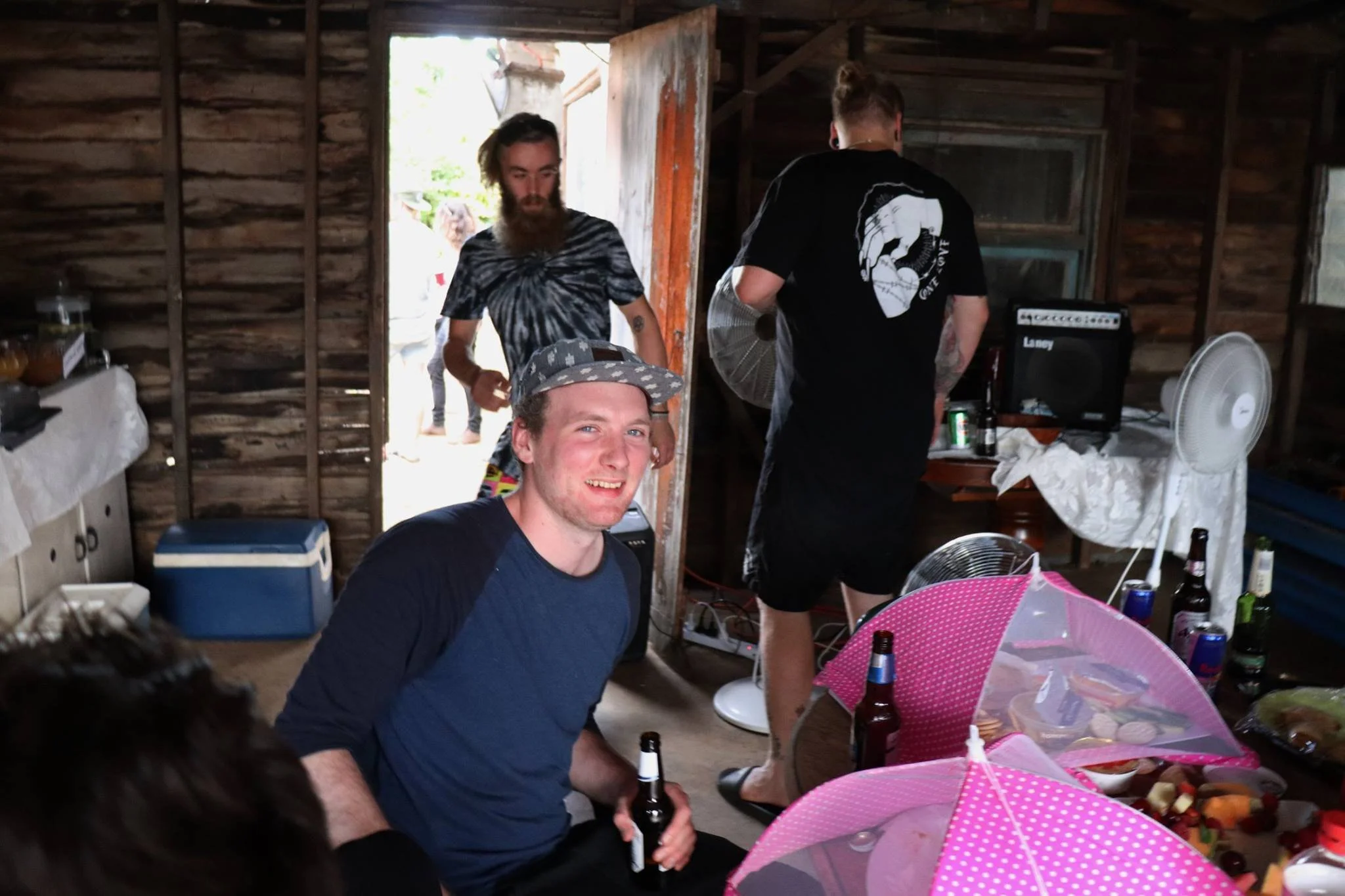 People gathered in a rustic indoor space, some holding beer bottles, with a man smiling at the camera in the foreground and others preparing food or music equipment, surrounded by tables and umbrellas.