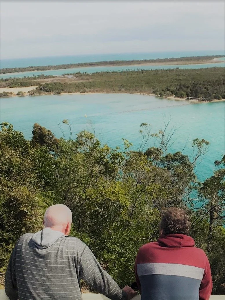 Two men sitting on a hill overlooking a bay with turquoise water, surrounded by greenery, with more water and land in the distance under a cloudy sky.