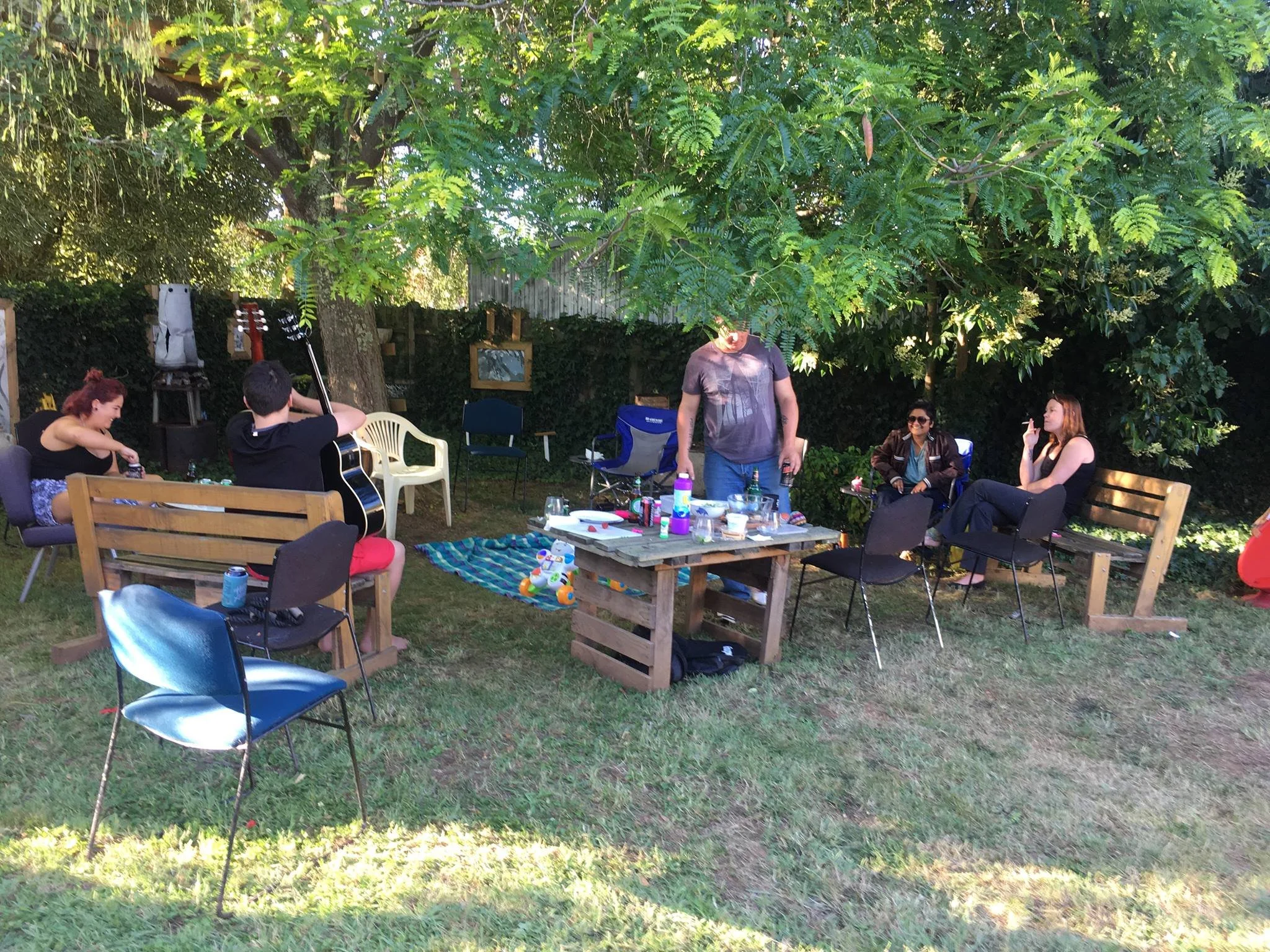 A backyard gathering with five people, including two people with guitars, sitting on chairs and benches under a large tree, with a picnic table, drinks, snacks, and decorations.