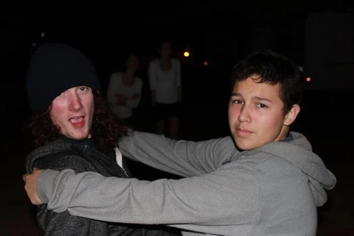 Two teenagers, one in a beanie and the other in a gray hoodie, embrace at night; the person on the left has curly hair and a slightly open mouth, while the person on the right looks at the camera with a neutral expression.