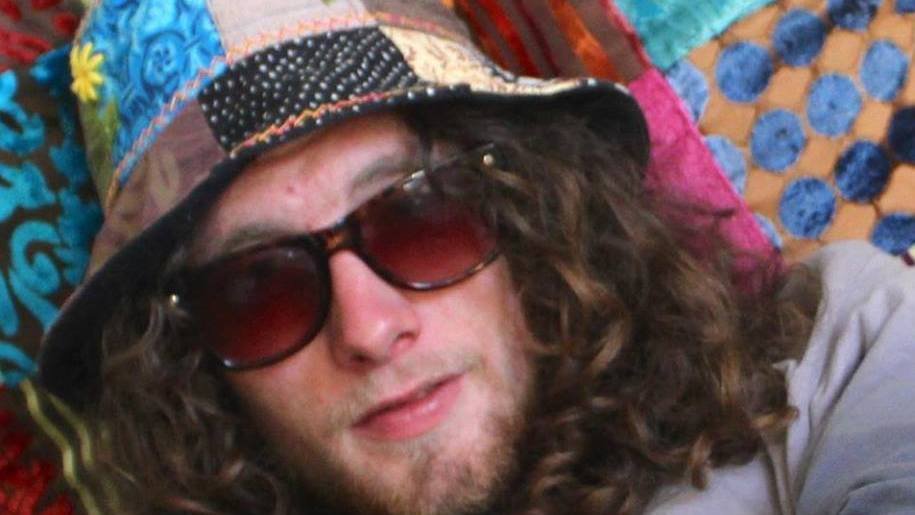 Person with long curly hair wearing a colorful patchwork hat, dark sunglasses, and a light-colored shirt, resting on a multicolored quilt.