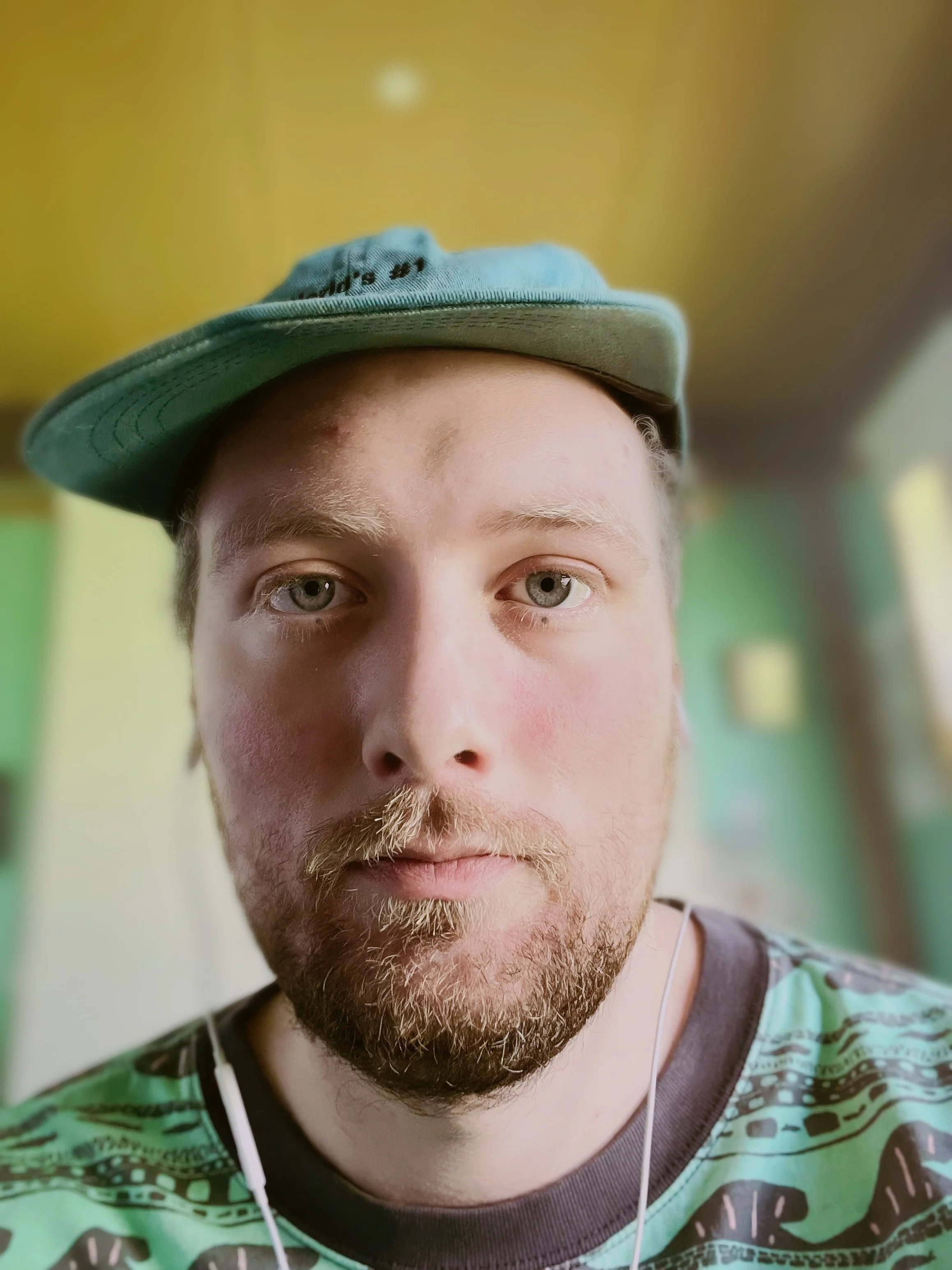 A young man with light-colored hair, light beard, and light eyes wearing a blue hat tilted to the side and a colorful, patterned shirt, looking directly at the camera with a blurred background.