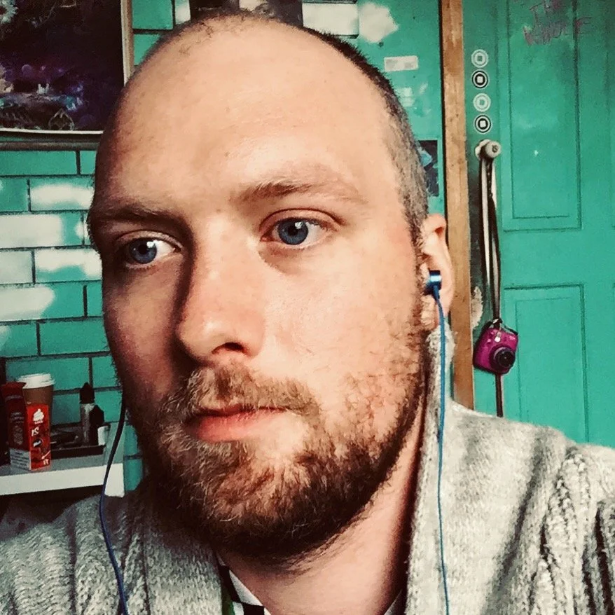 Close-up of a man with blue eyes, light skin, a beard, and a mustache, wearing earphones, in front of teal-colored lockers and a brick wall, with a camera hanging on the locker.