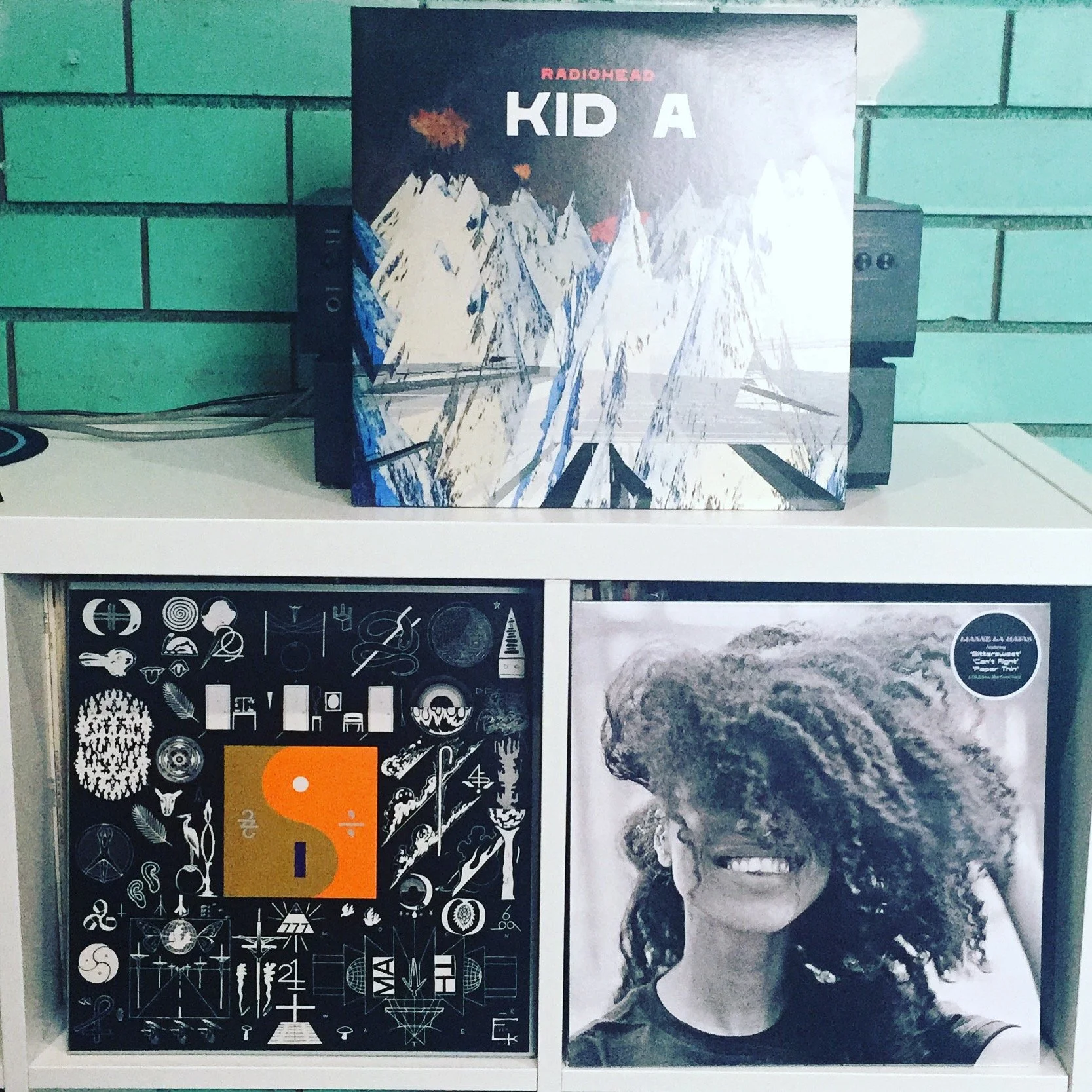 Three record albums displayed on a shelf, featuring a Radiohead Kid A album, a black and white album with various small icons including a woman, a crescent moon, and a leaf, and a black-and-white photo of a woman with curly hair smiling.