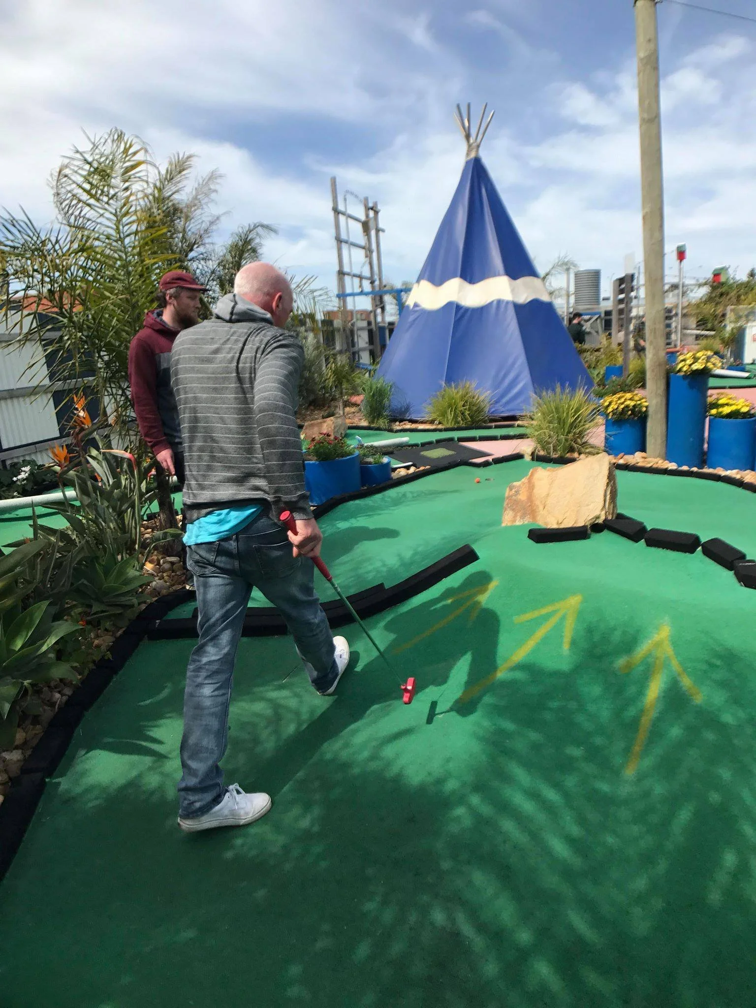 Two men playing miniature golf outdoors, one hitting the ball with a club while the other watches, with a teepee-shaped structure and various plants and potted flowers in the background.