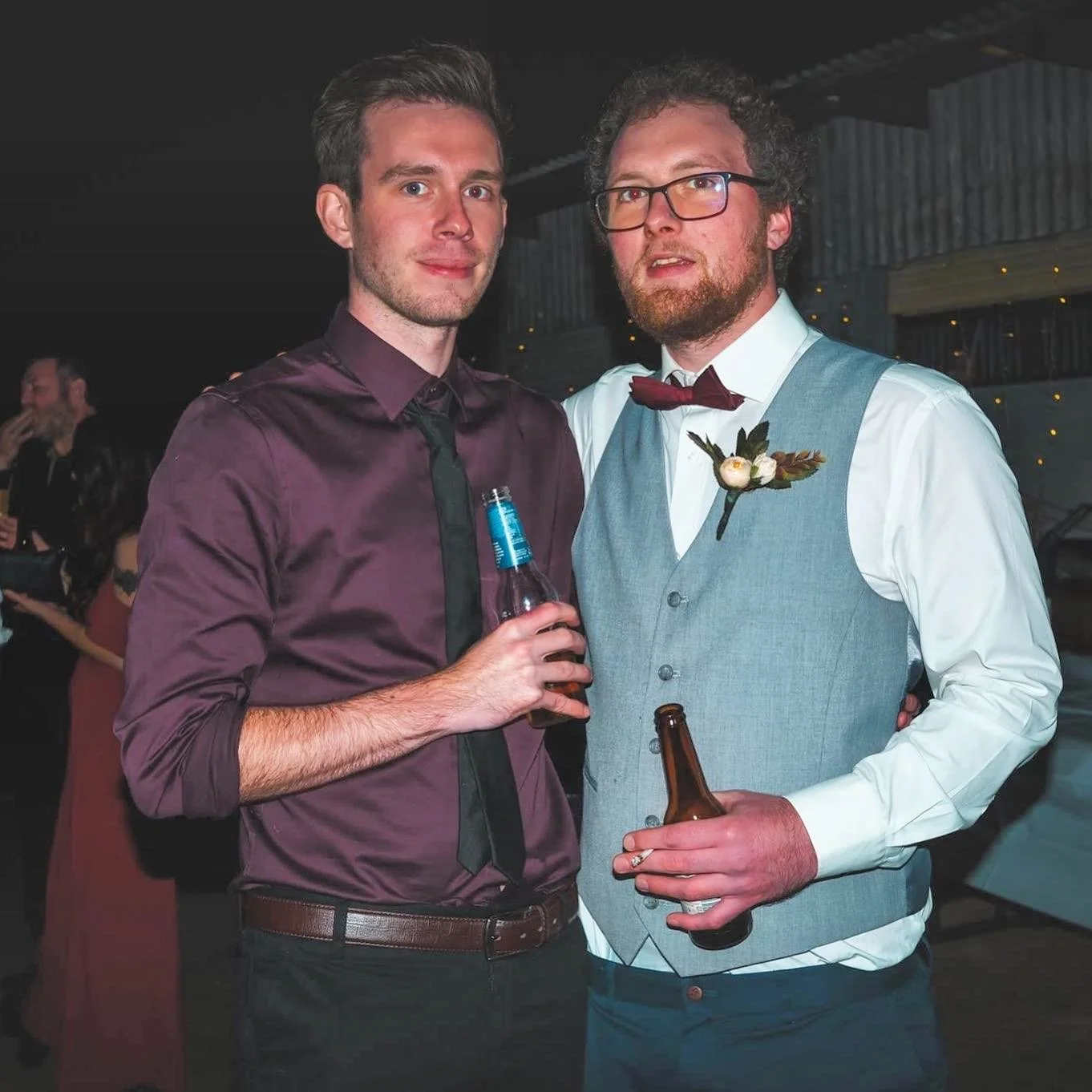 Two young men at a social event holding bottles of beer, dressed in formal attire, standing close together with a dark indoor background.