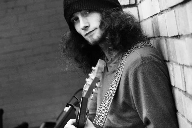 A young man with long curly hair, wearing a beanie, leaning against a brick wall and holding an acoustic guitar.